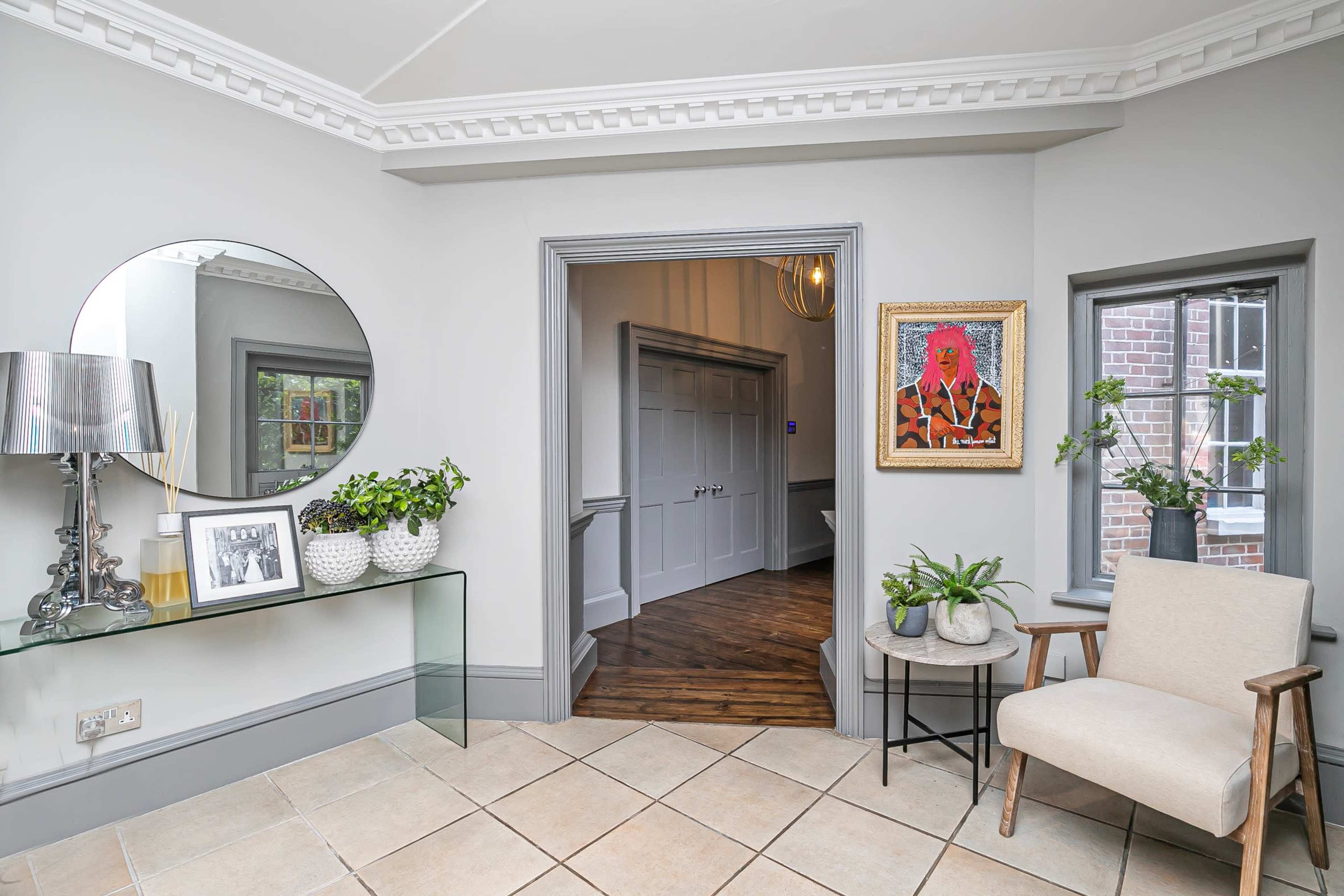 A modern entryway features a circular mirror, a console table with decorative items, a chair, and a doorway leading to a room with wooden flooring.