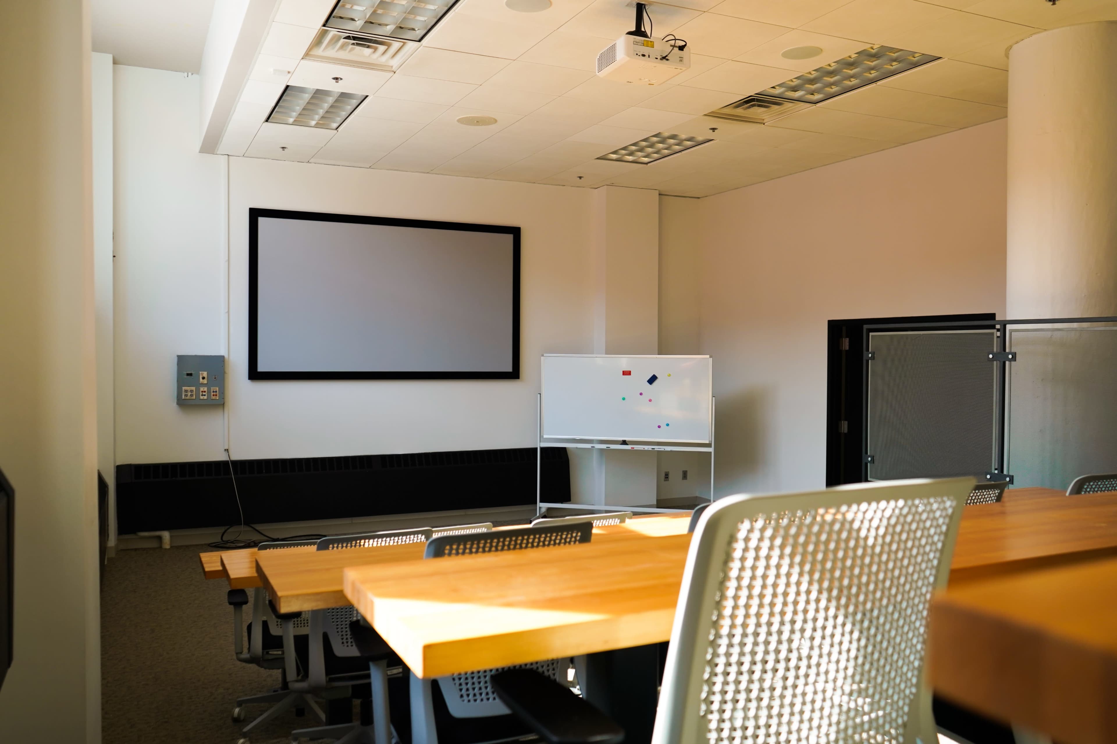 The image shows a modern conference room with a large screen, a whiteboard, and several tables facing the screen.