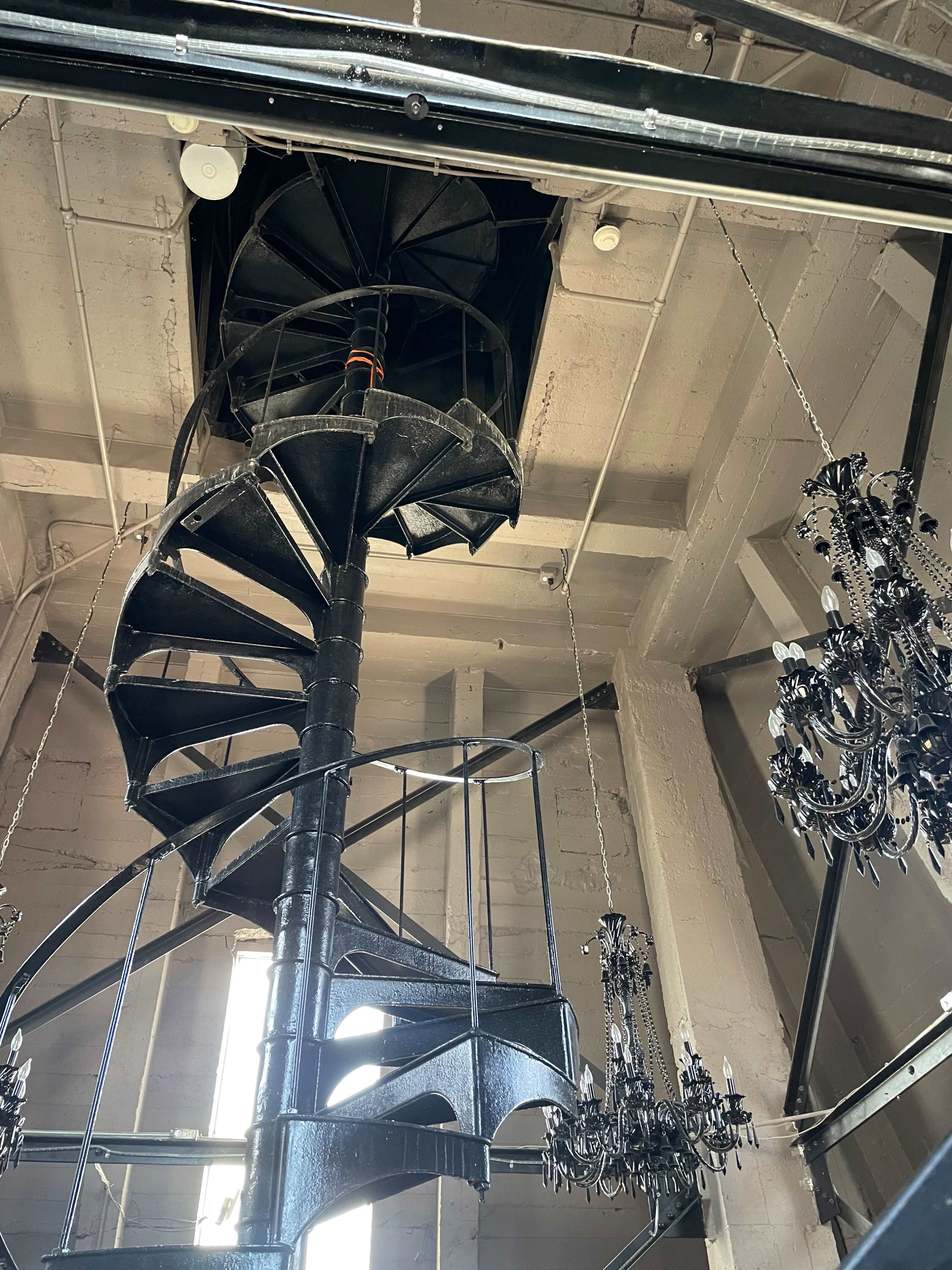 A spiral staircase with black metal steps leads up to a ceiling opening, surrounded by decorative chandeliers.