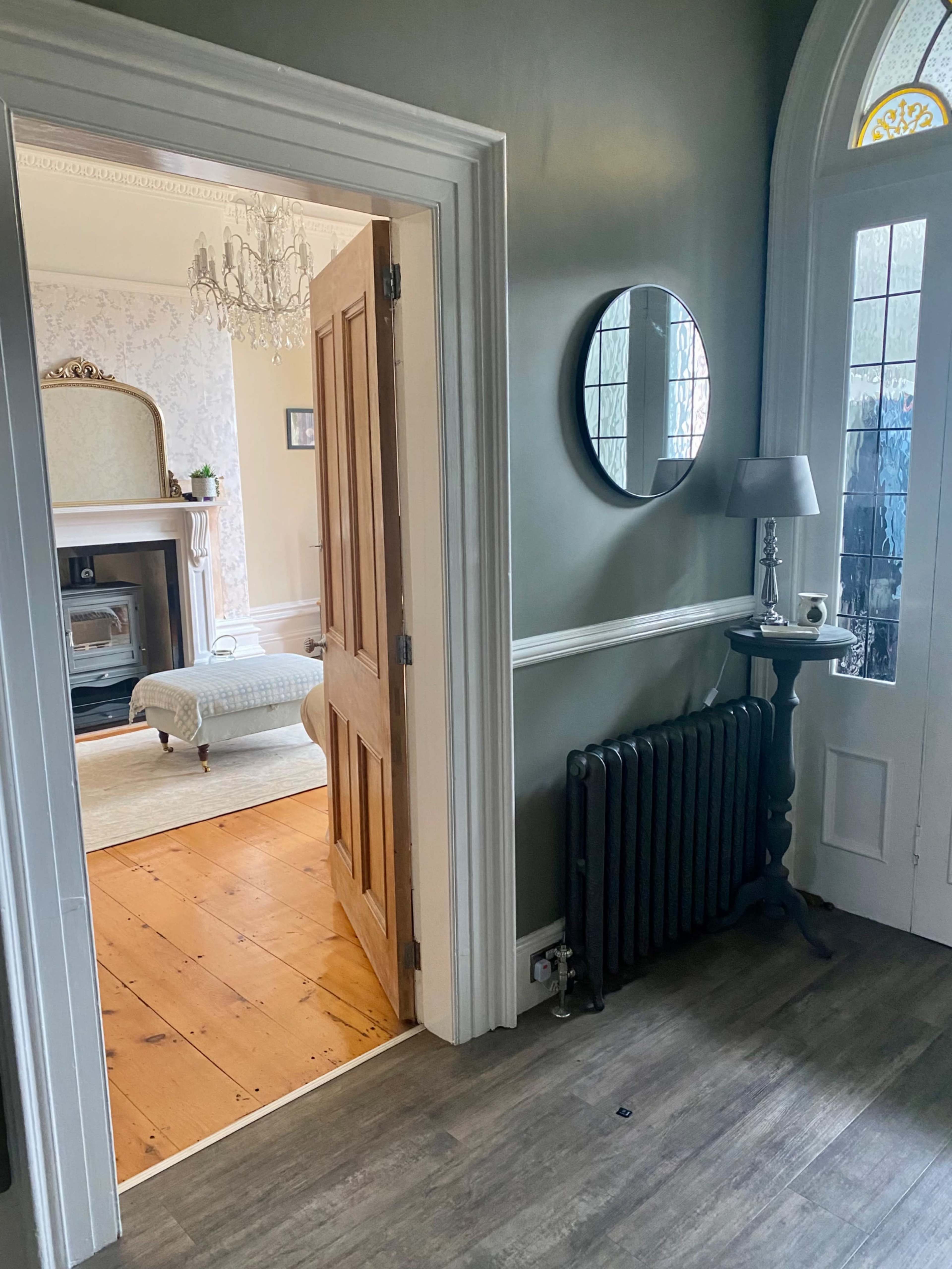 A hallway with an open wooden door leading into a room featuring a fireplace, a mirror on the wall, and a radiator.