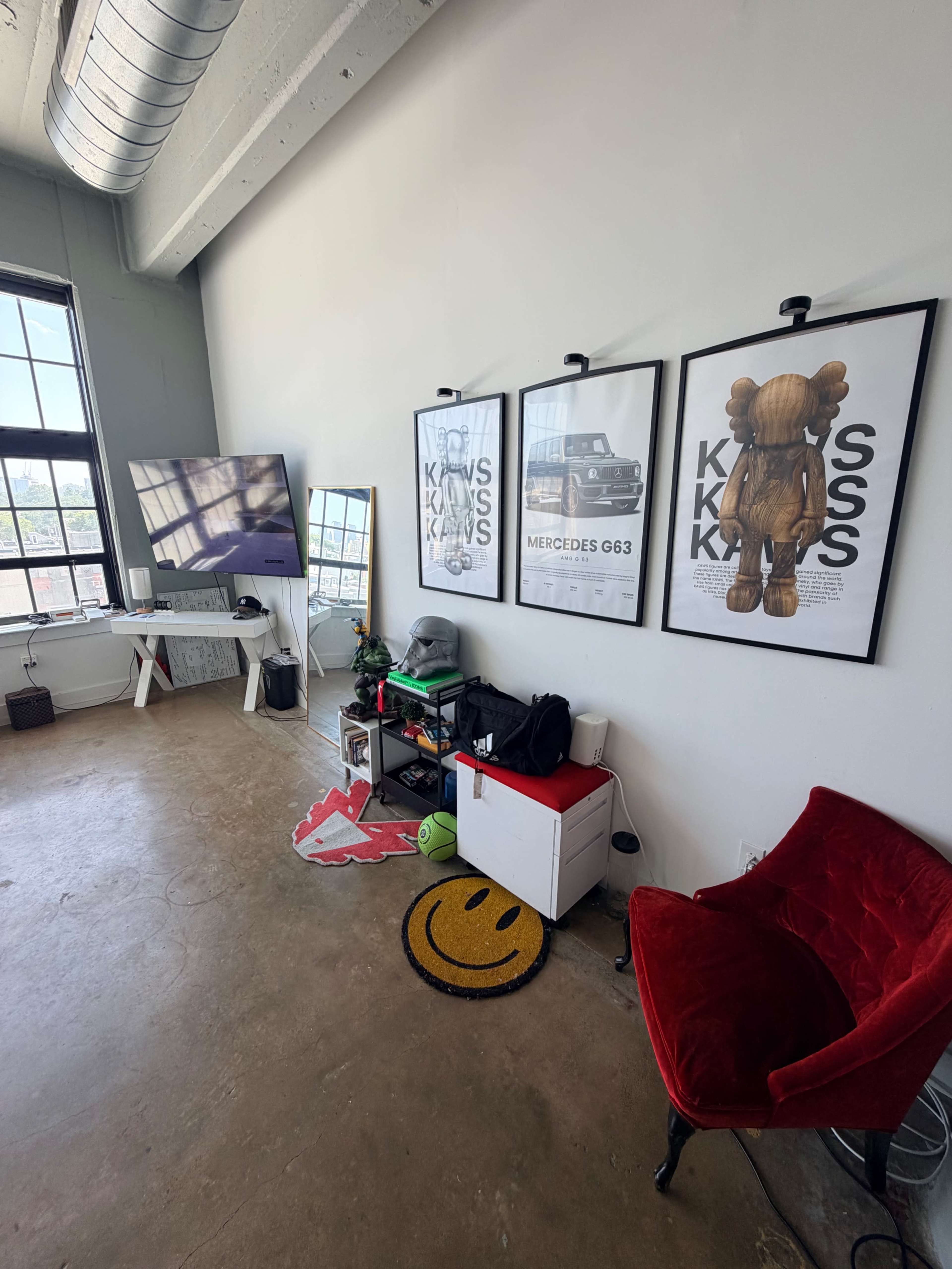 The image shows a modern room featuring a large window, a television mounted on the wall, and three framed posters, along with a red chair and various items on the floor.
