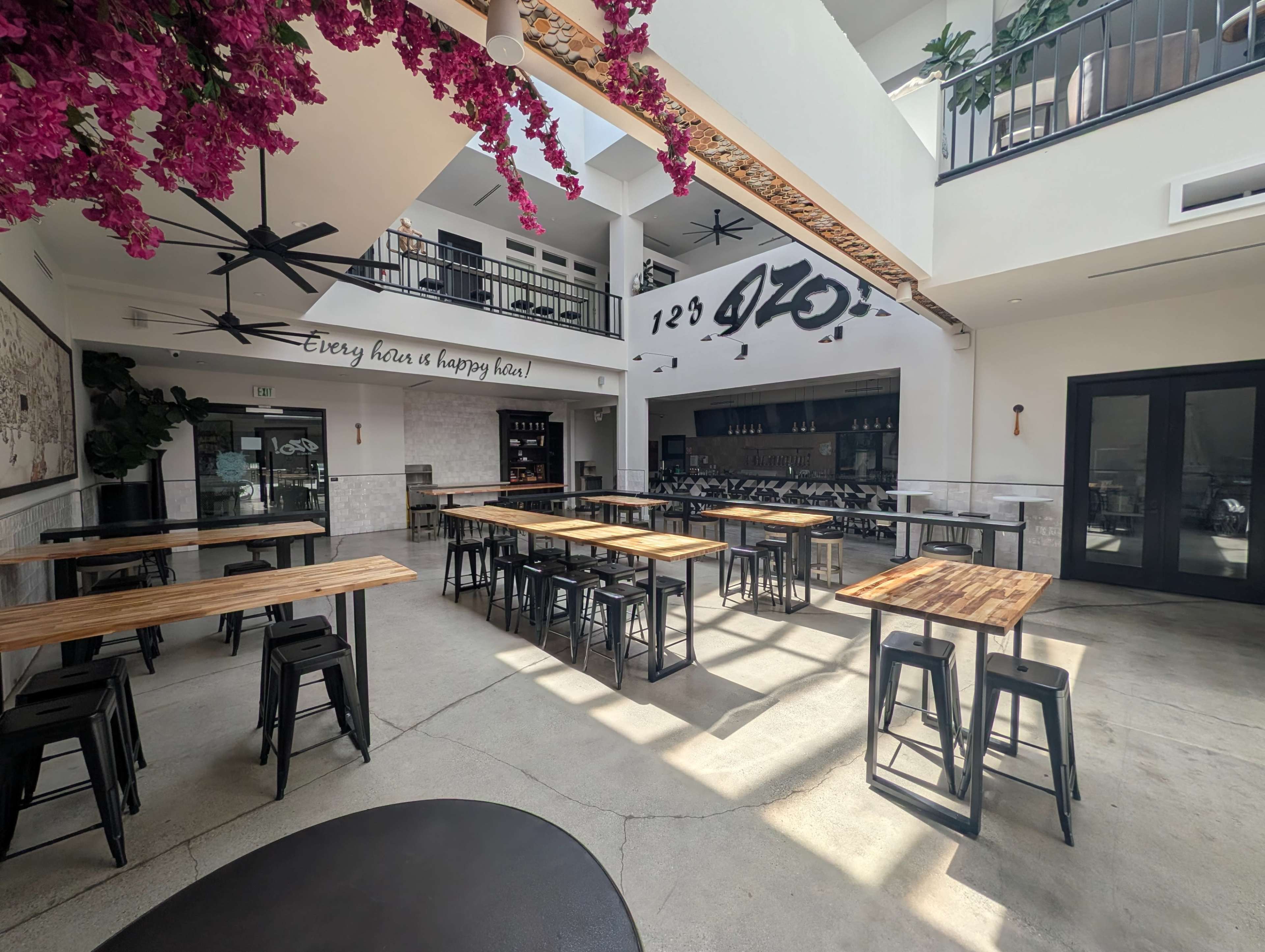 The image shows a spacious restaurant interior with wooden tables and black metal chairs, featuring large fans and a bar area with shelves behind it adorned with bottles.