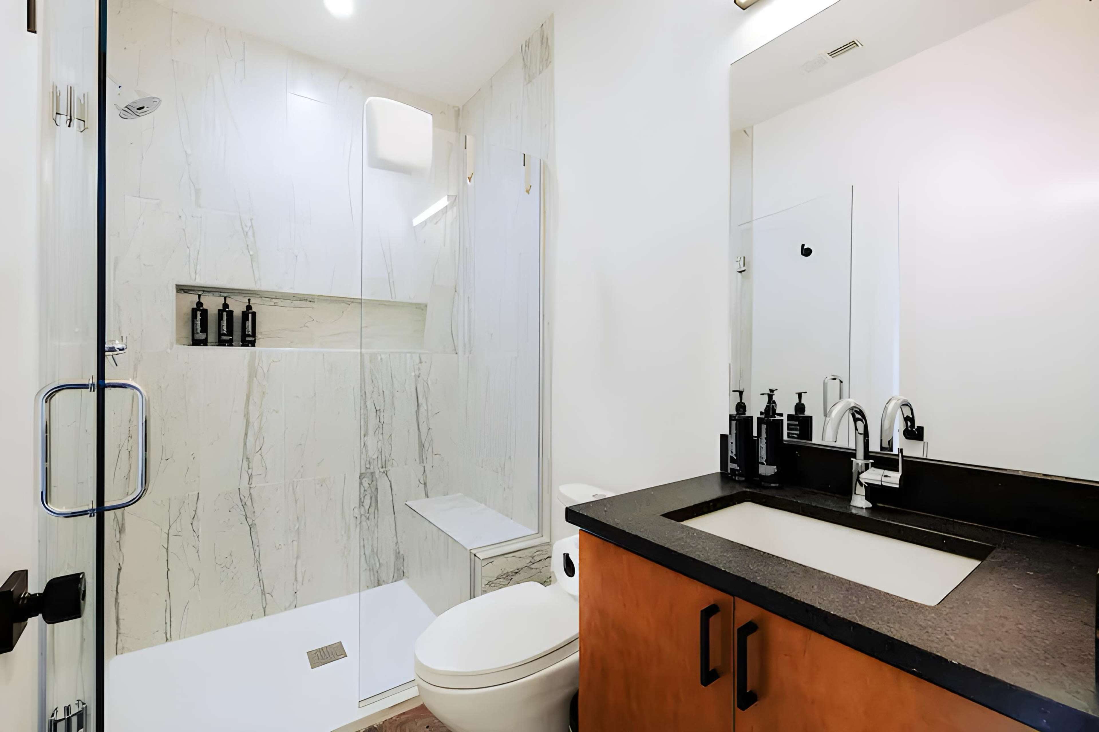 The image shows a modern bathroom featuring a glass shower, marble walls, a sleek vanity with a dark countertop, and organized toiletries.