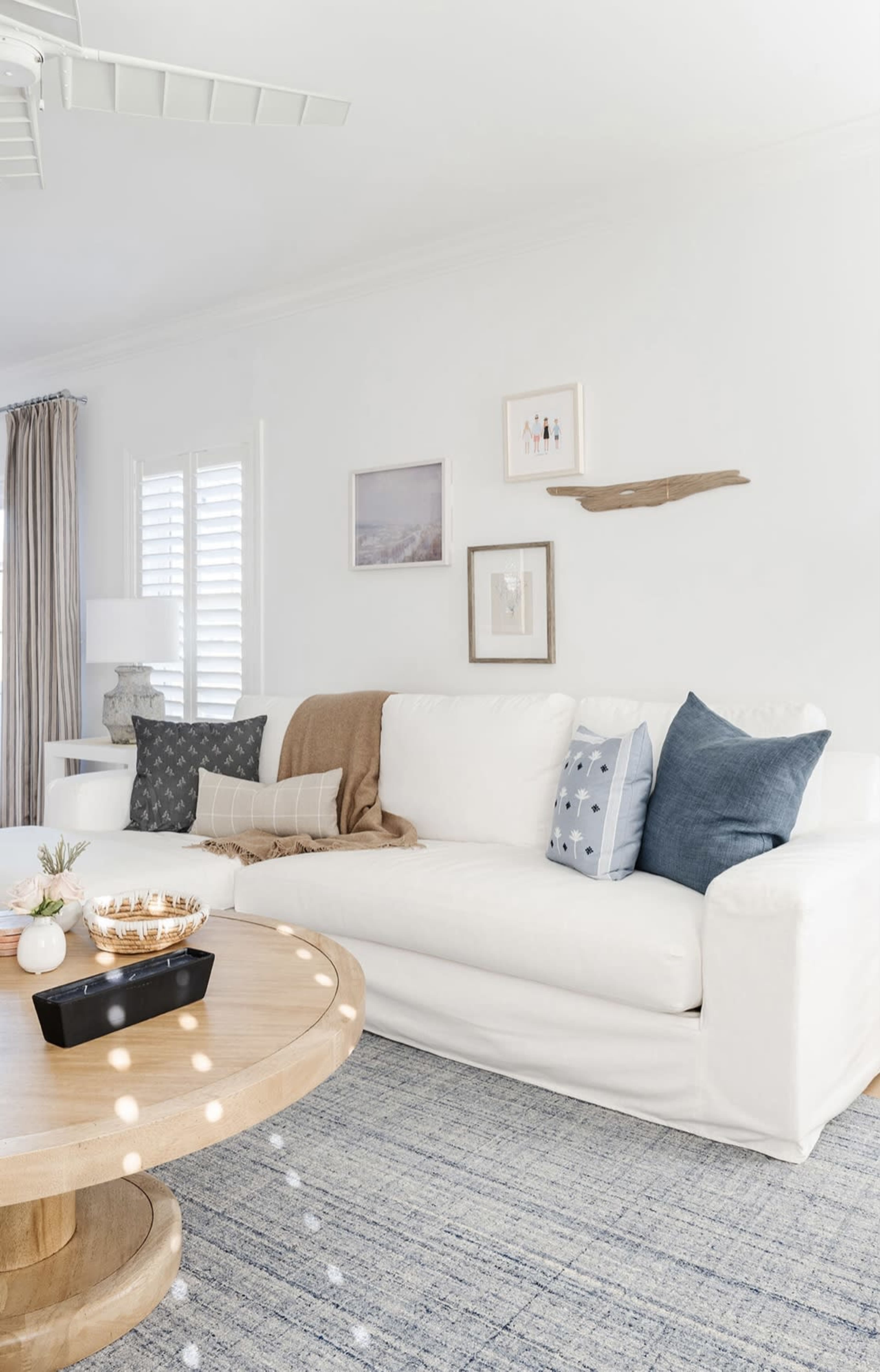 A modern living room features a white sofa with decorative pillows, a wooden coffee table, and a light-colored rug, complemented by natural light and minimal decor.