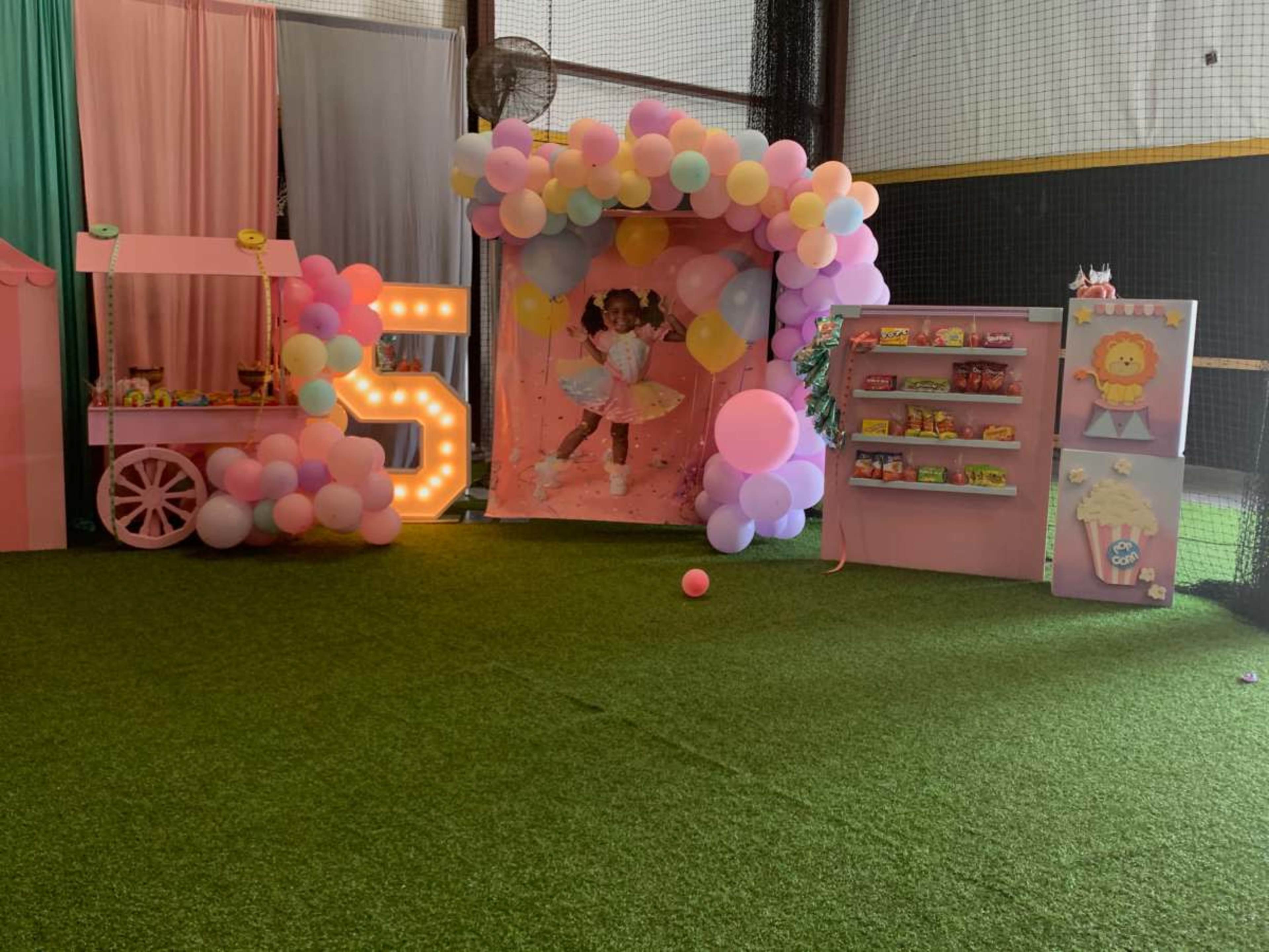The image shows a decorated indoor party area featuring a balloon arch, a photo backdrop, a candy cart, and a shelf with snacks on a faux grass floor.