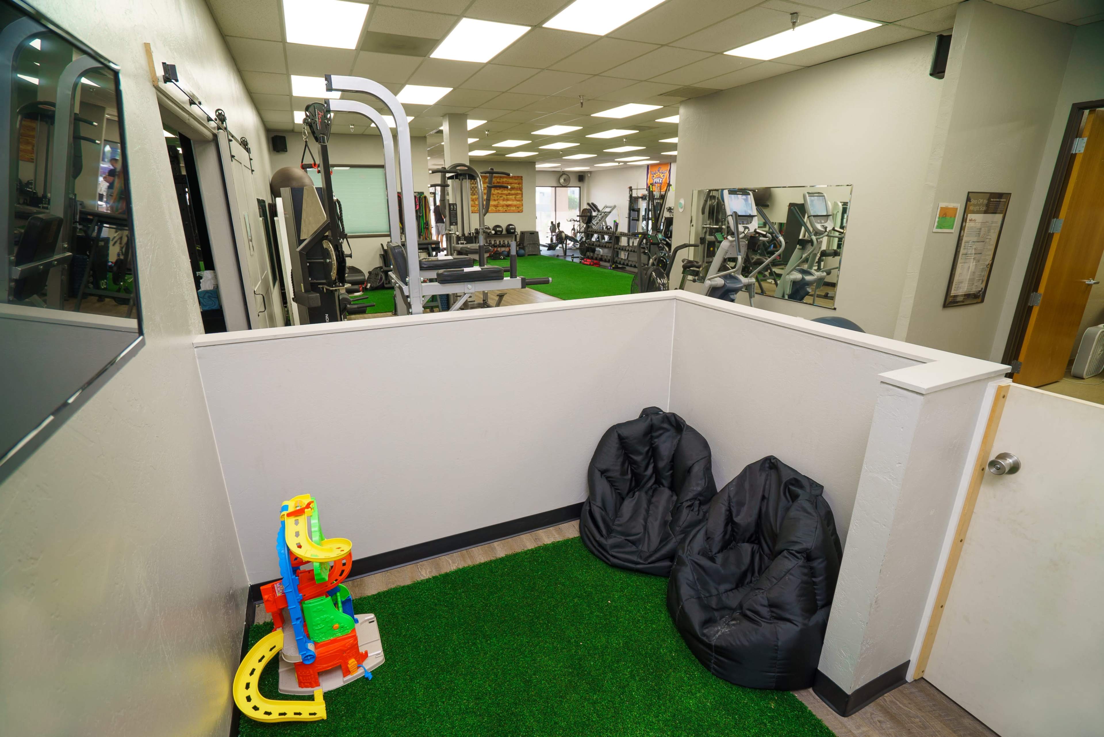 The image shows a fitness facility featuring workout equipment, mirrors, and a small play area with colorful toys and bean bags in a corner.