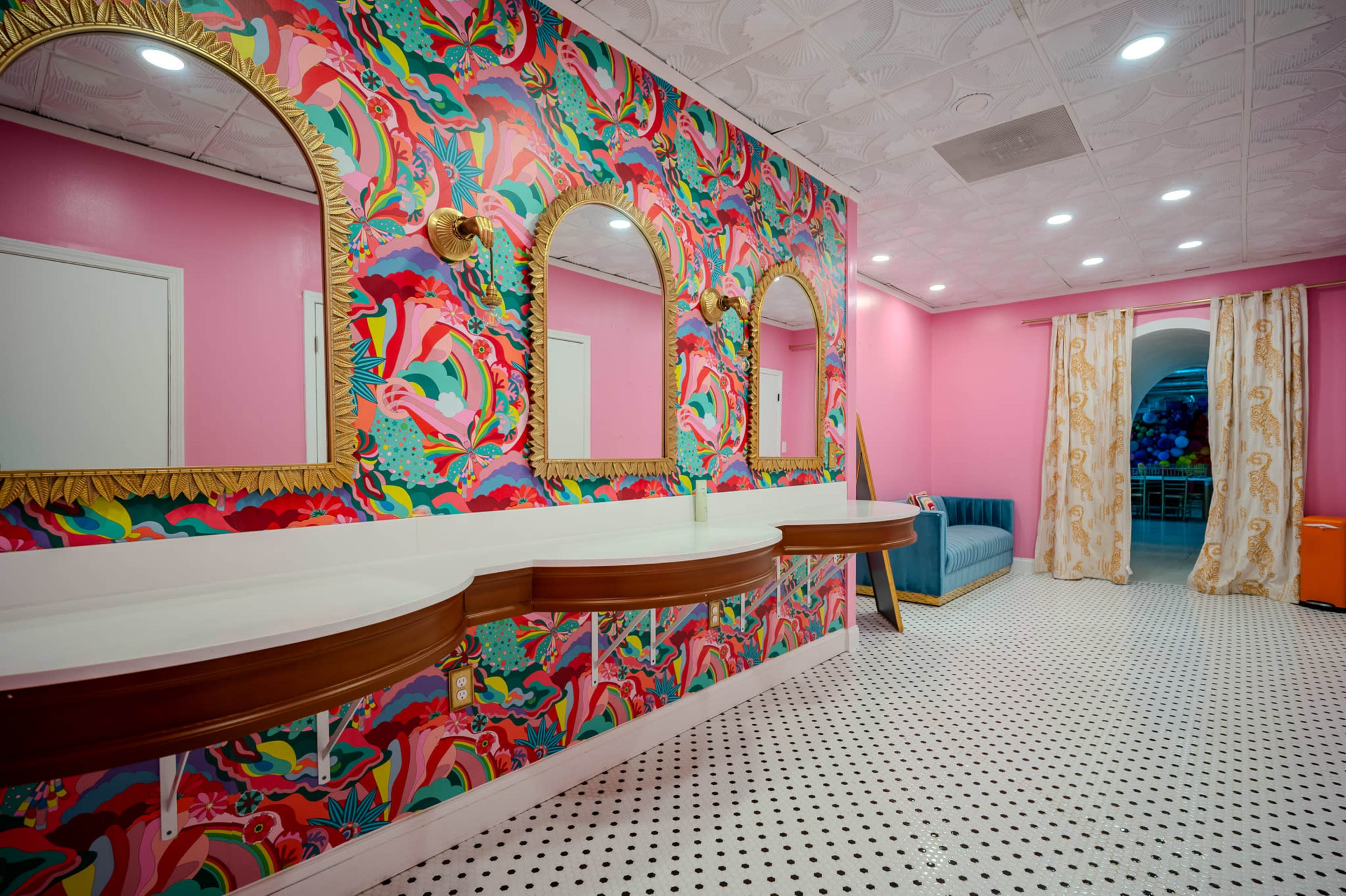 The image shows a brightly colored bathroom with vibrant floral-patterned wallpaper, three round mirrors, and a white countertop, featuring a seating area with a blue couch and patterned curtains.