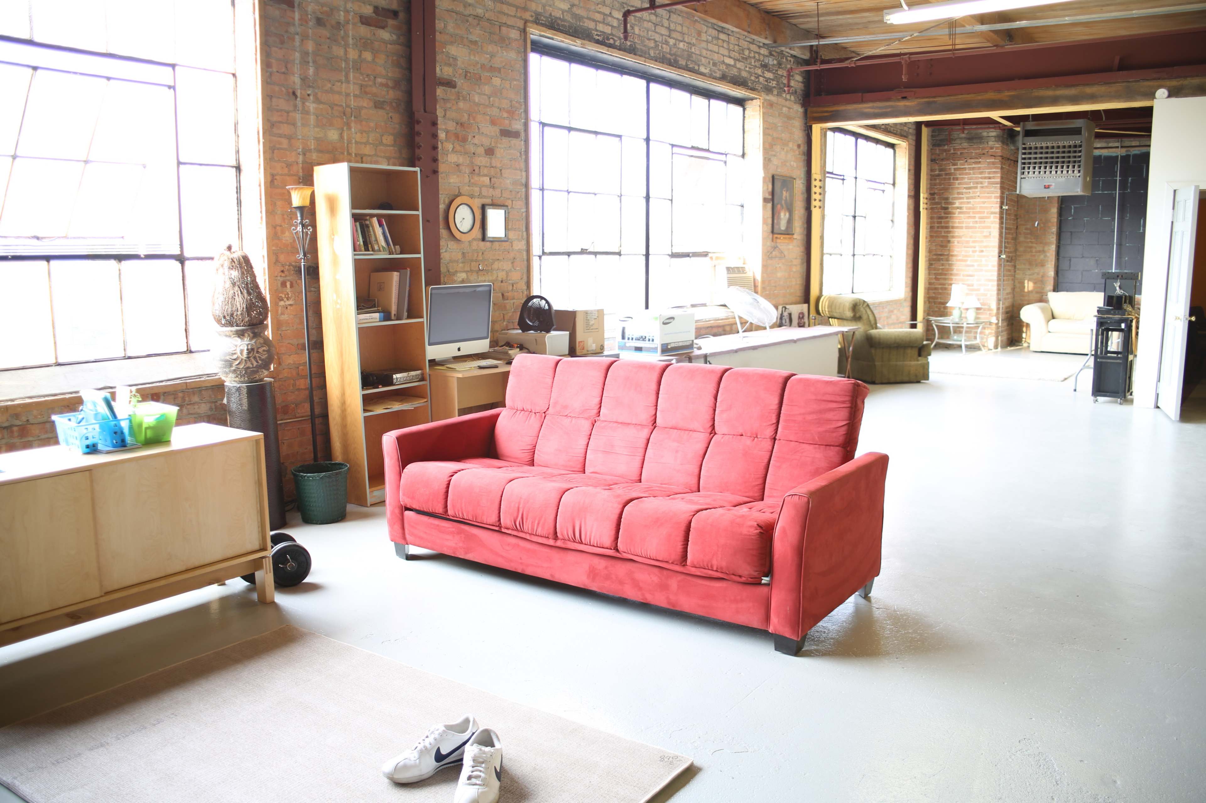 A spacious, industrial-style loft with large windows, featuring a red sofa in the foreground and various pieces of furniture in the background.