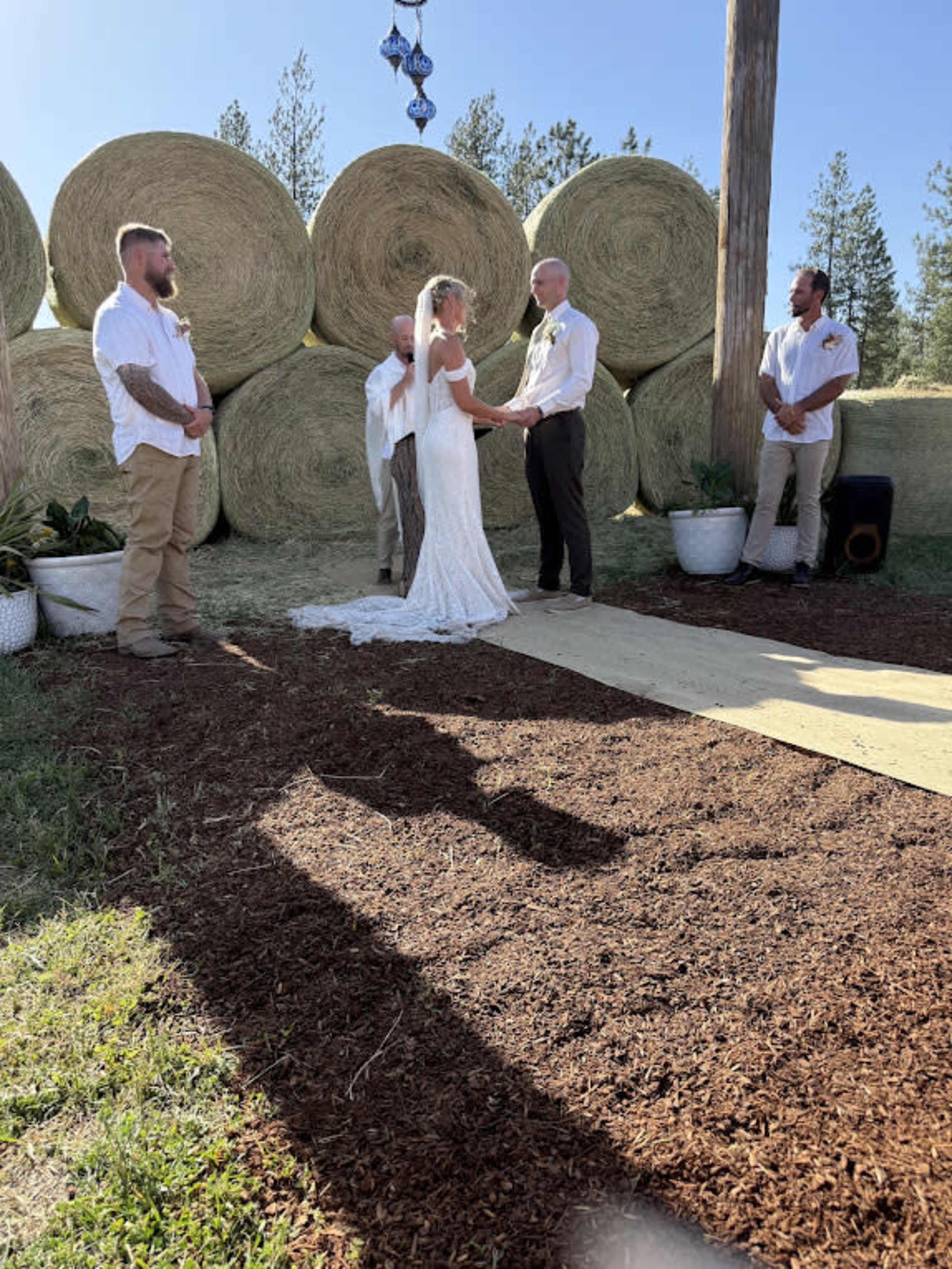 A couple stands on a wooden platform exchanging vows in front of a small gathering, with large hay bales in the background.