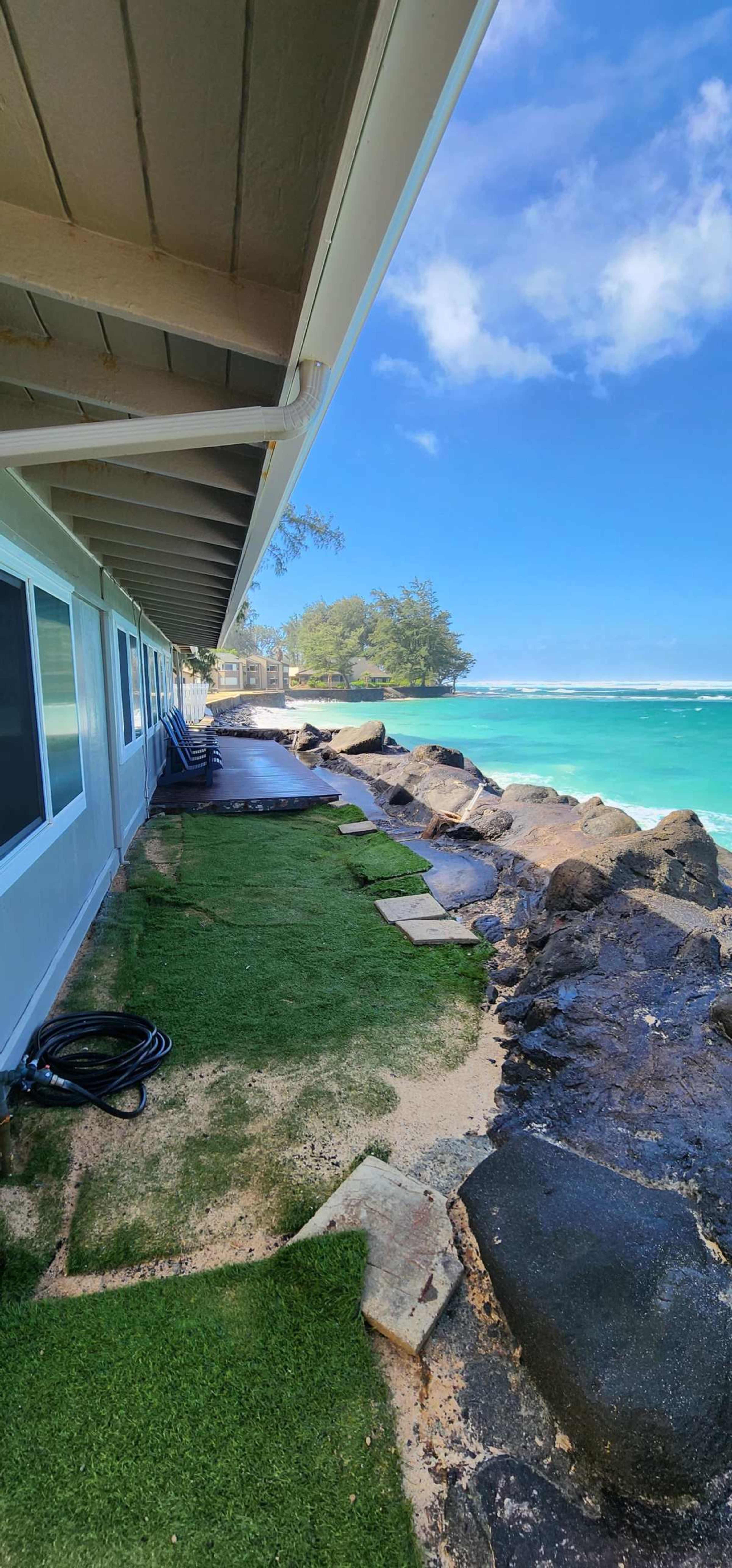 A pathway lined with artificial grass runs along a coastline next to a building, with waves gently lapping against rocky shorelines under a clear blue sky.