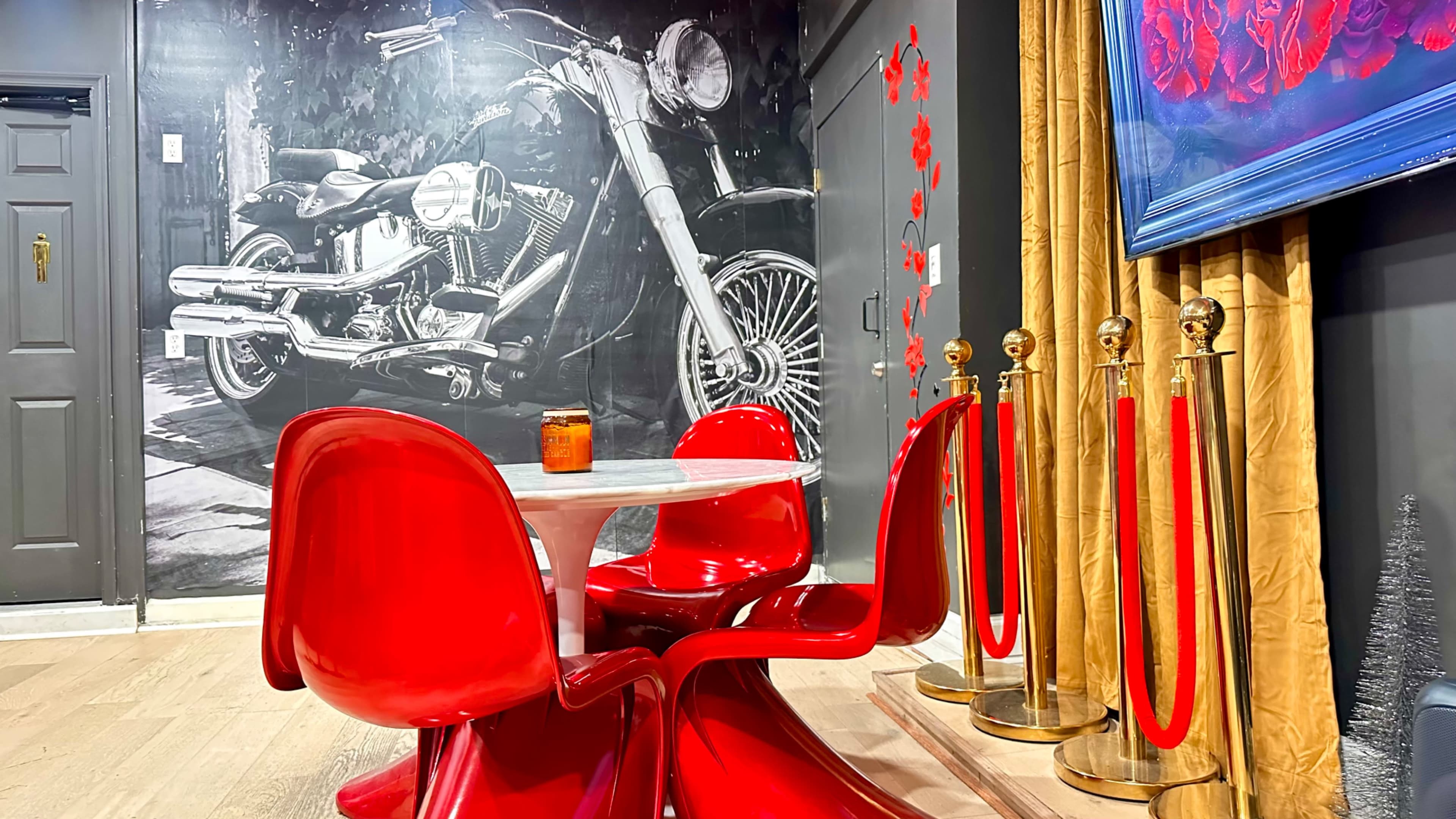 A modern dining area features a round table surrounded by red chairs, with a motorcycle mural on the wall and a velvet curtain nearby.