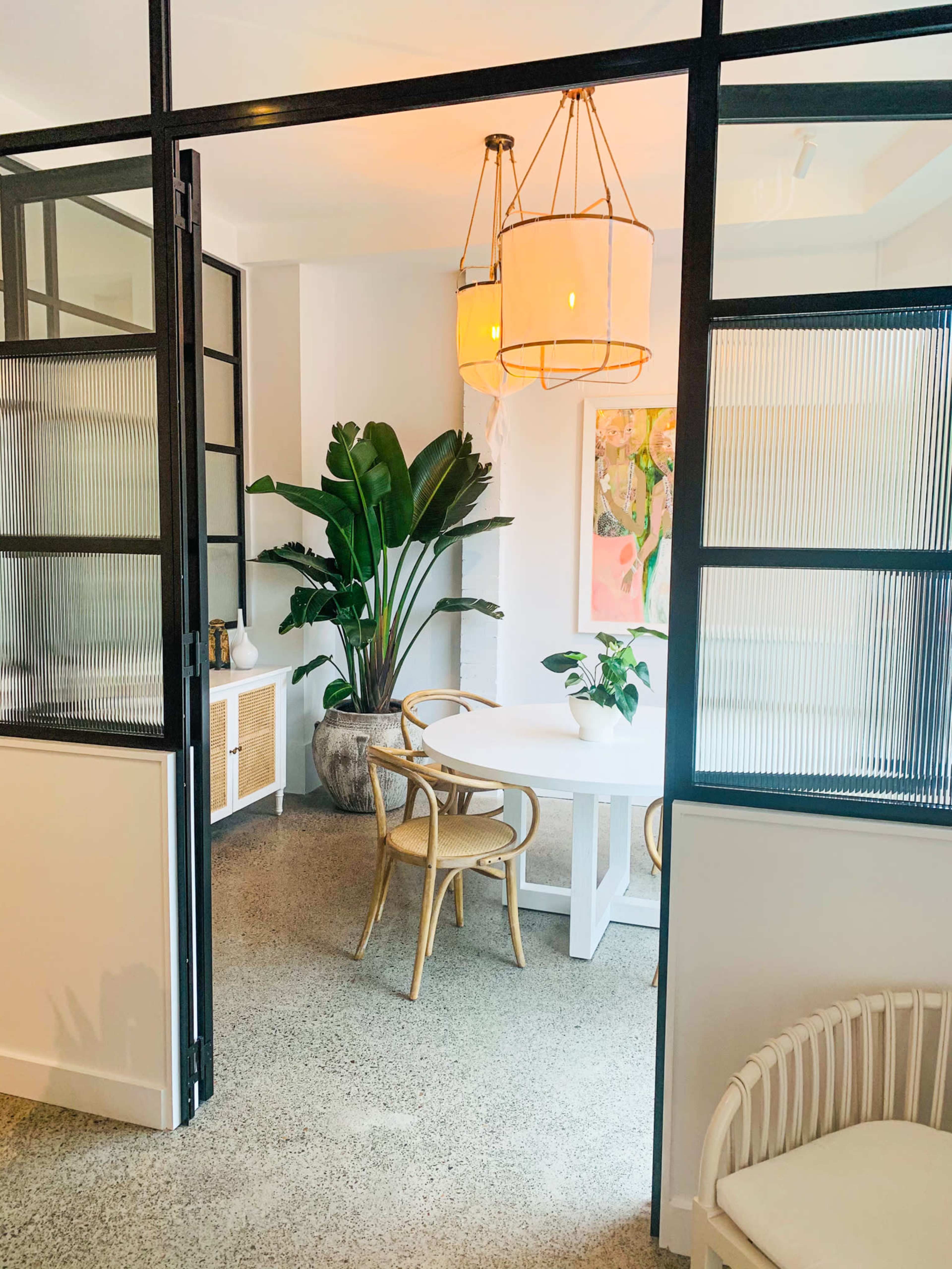A bright, modern dining area features a round table surrounded by chairs, with large plants and decorative art displayed on the walls.