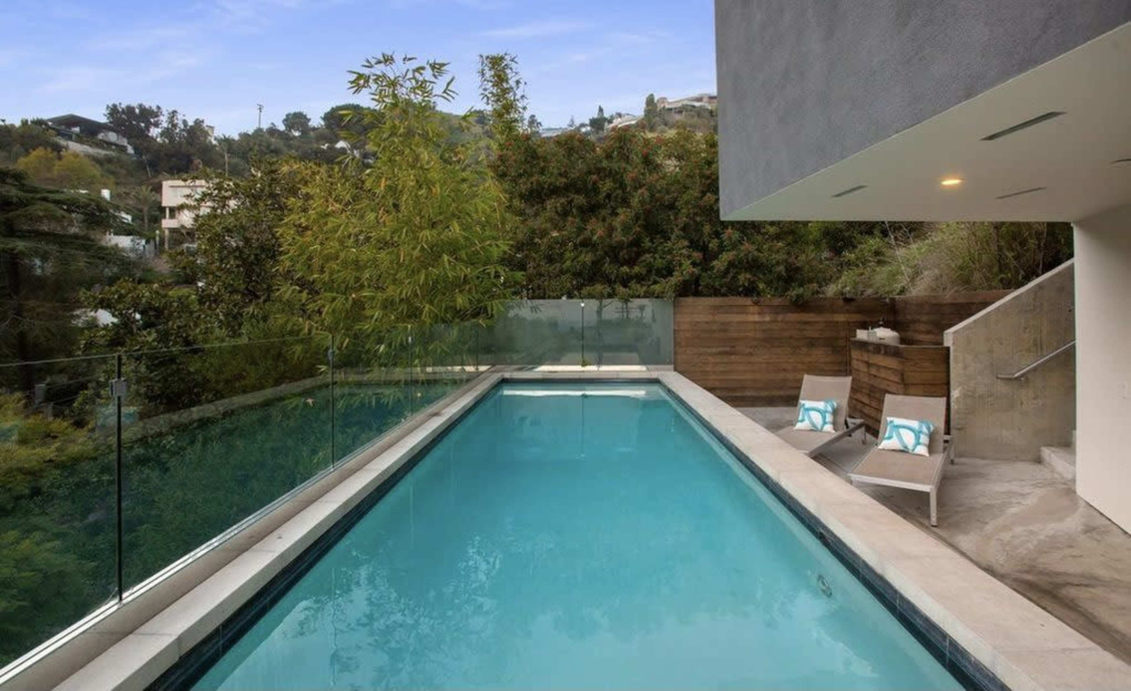 The image shows a modern swimming pool surrounded by greenery and contemporary outdoor furniture.