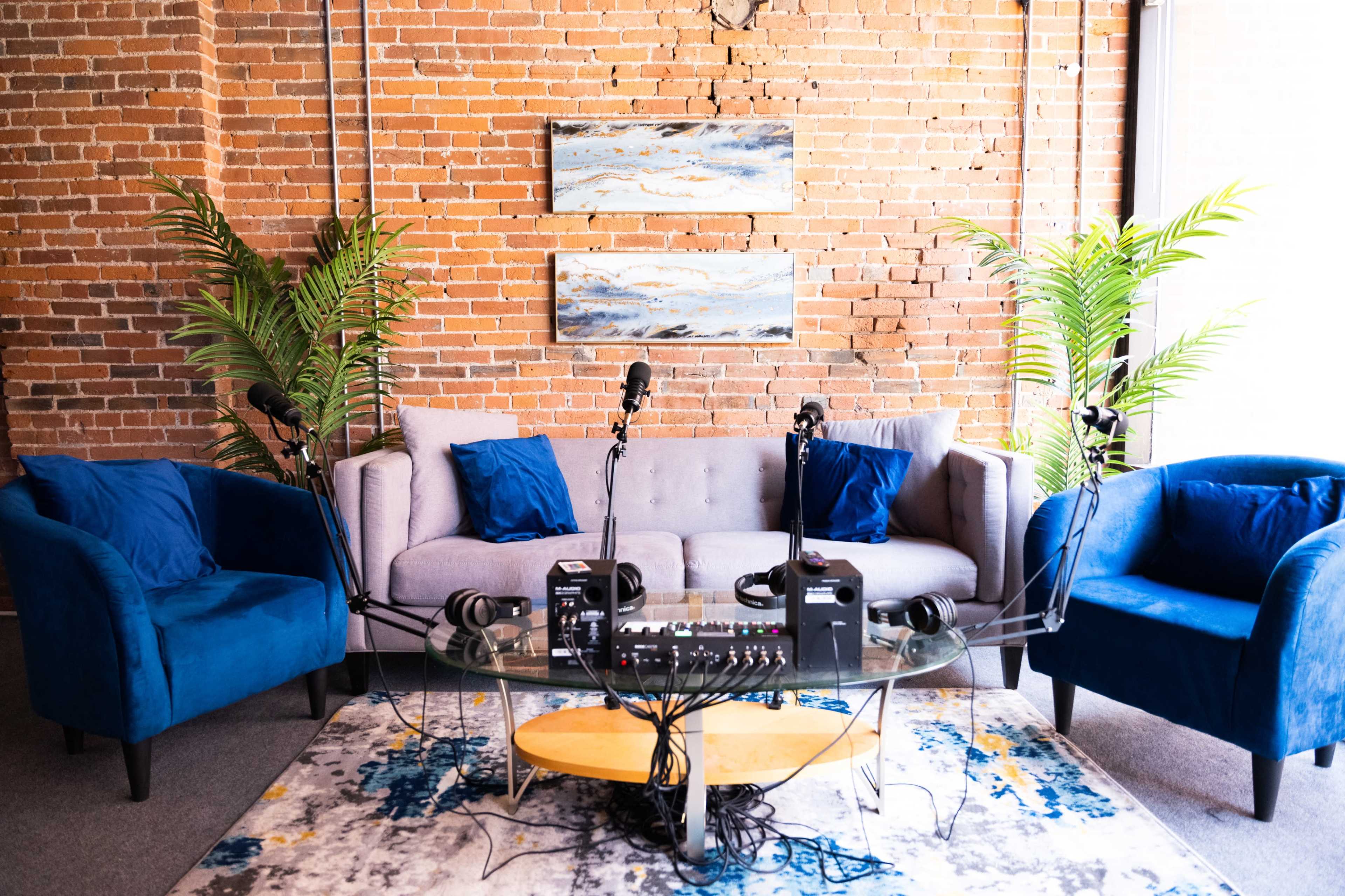 The scene shows a modern podcasting setup with a gray couch and blue chairs, featuring microphones and headphones on a glass table in front of a brick wall.