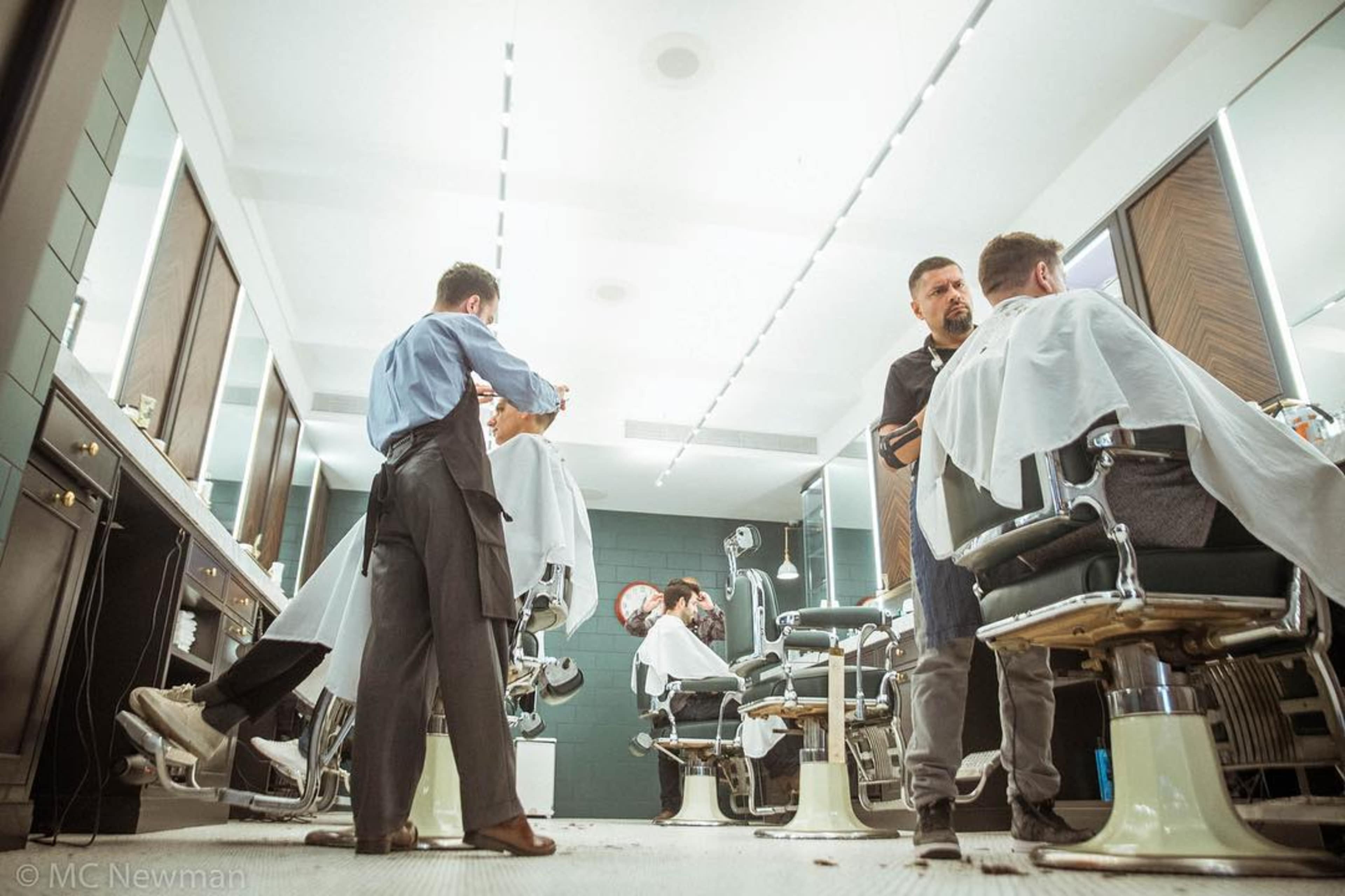 Two barbers attend to clients in a modern barbershop with stylish barber chairs and bright lighting.