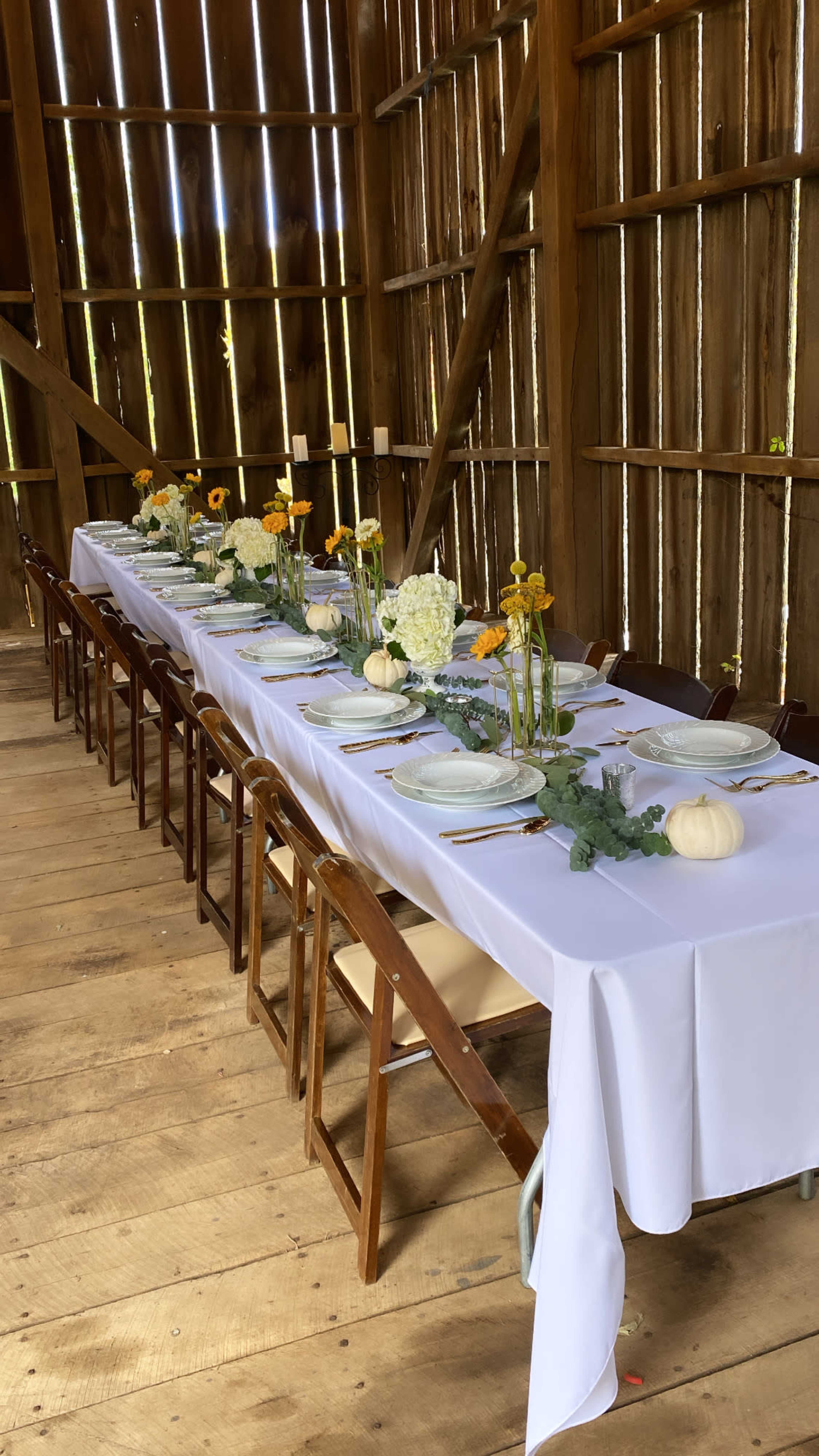 A long banquet table is set with white tablecloths, plates, and floral centerpieces inside a wooden barn.