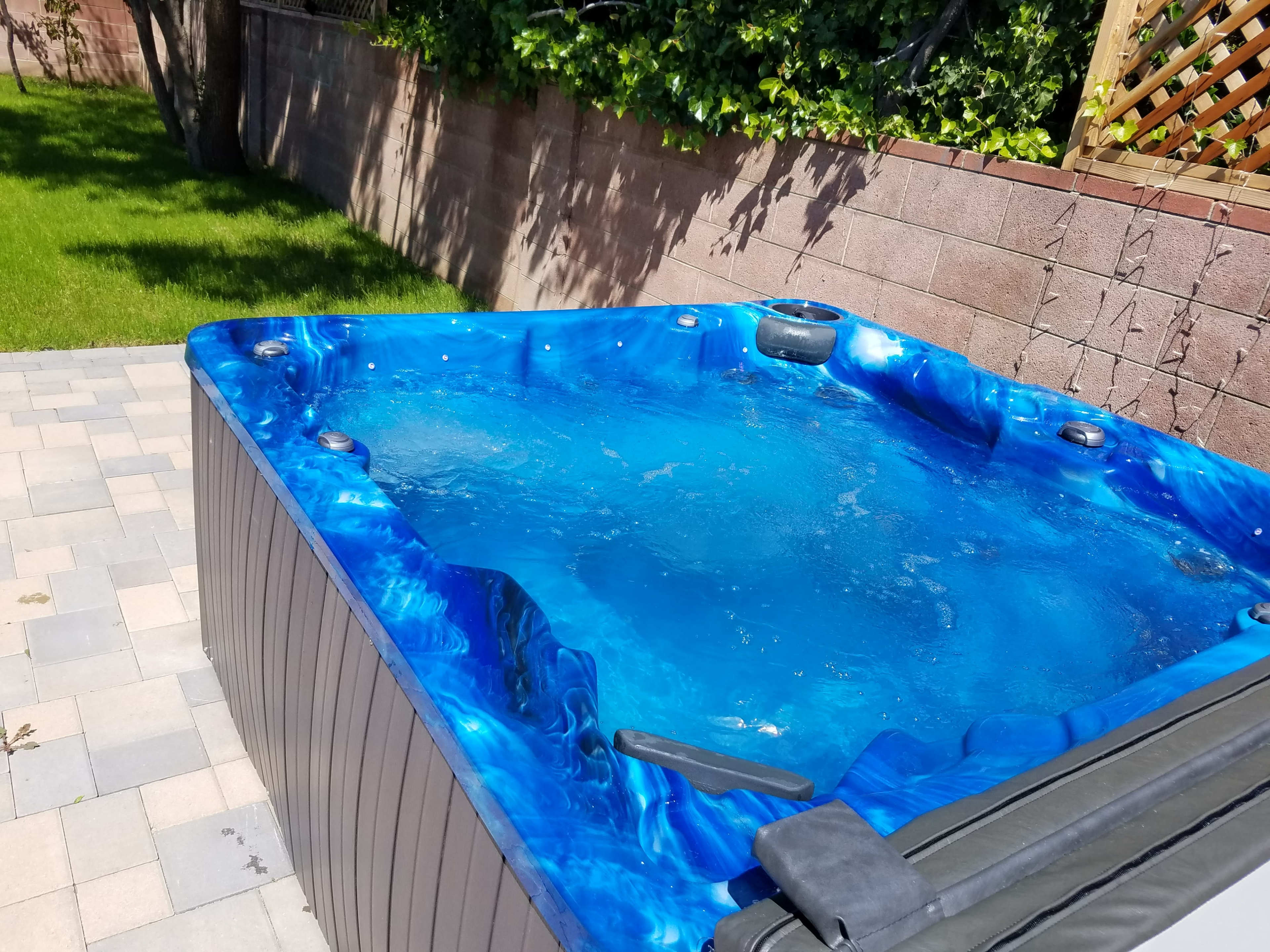A blue, triangular hot tub filled with water is situated in a yard with grass and a brick wall in the background.