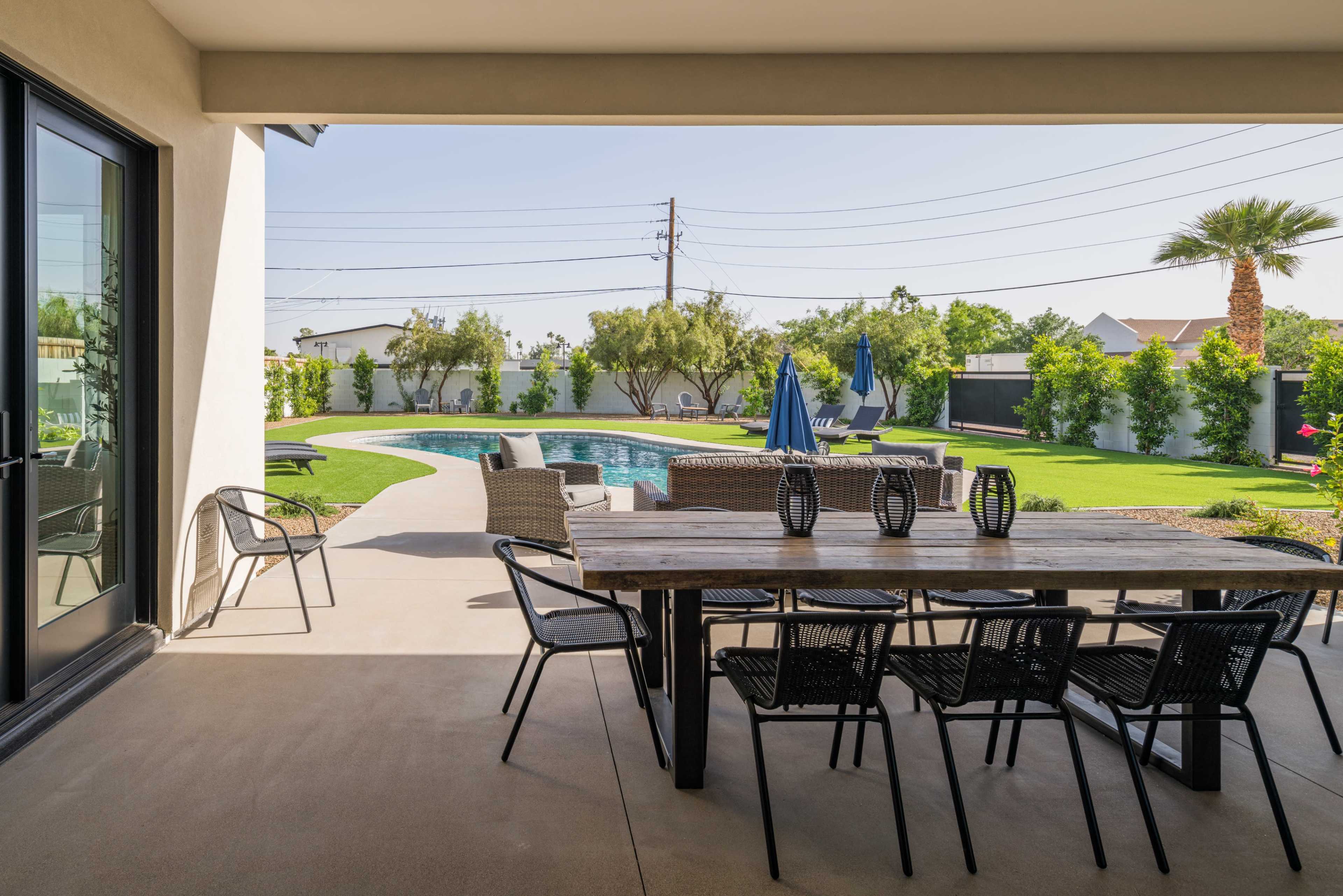 A patio area features a wooden dining table with chairs overlooking a pool surrounded by greenery and lounge chairs.