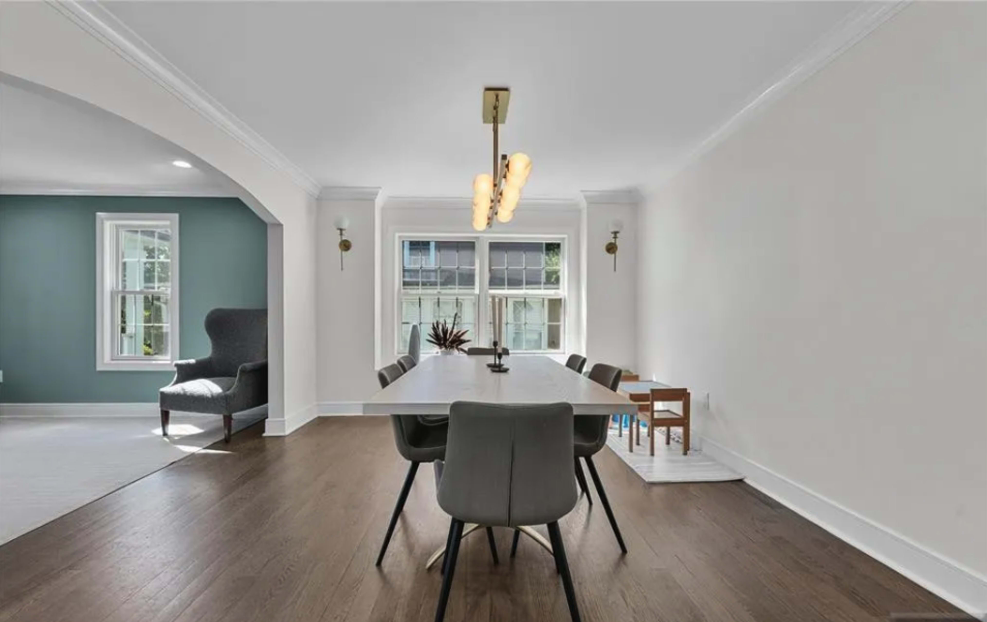 A dining area features a long table with modern chairs, illuminated by a contemporary light fixture, with large windows allowing natural light to fill the space.