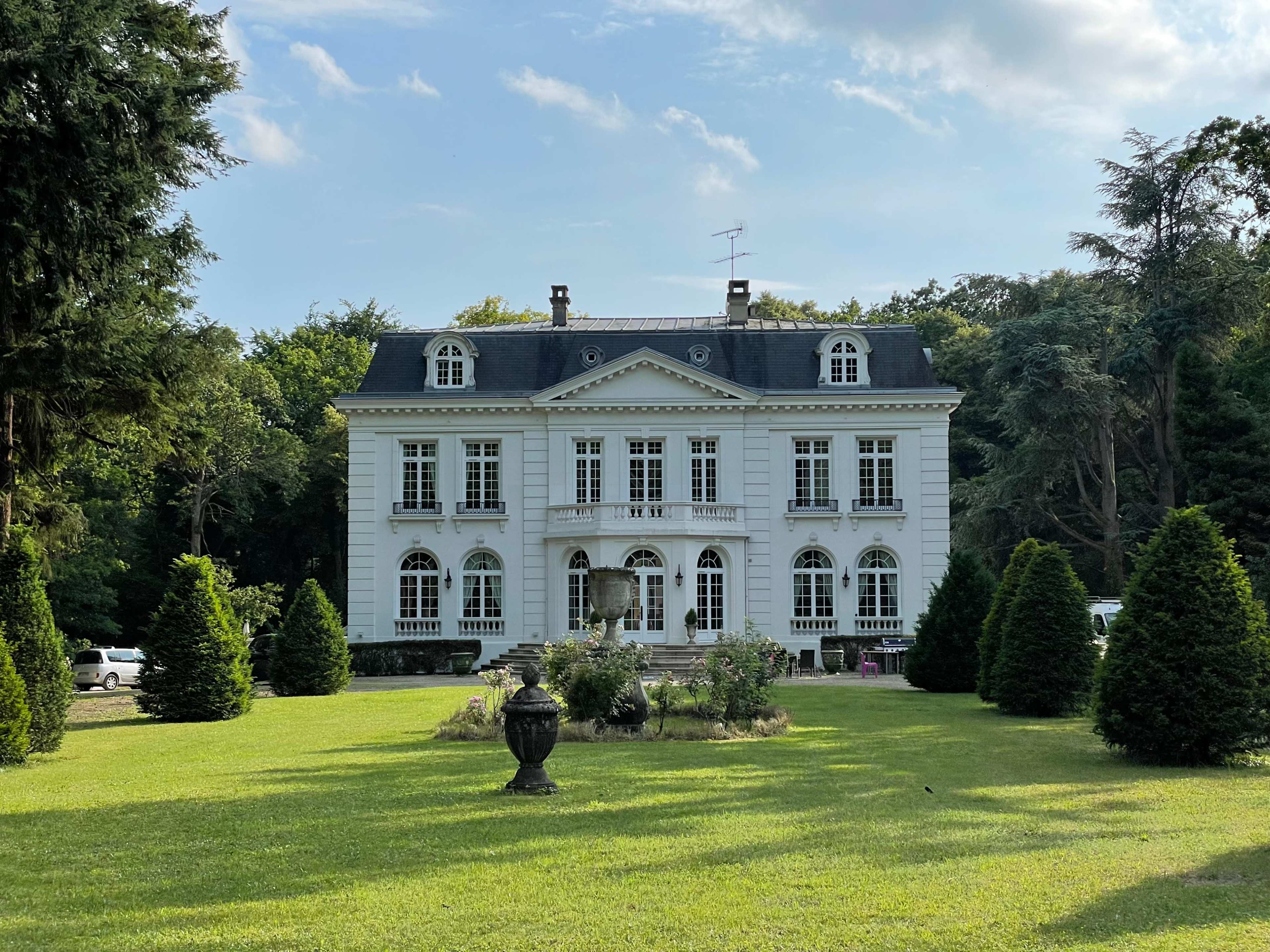 A large, gray mansion with multiple windows and a balcony is set on a green lawn surrounded by trees and neatly trimmed bushes.