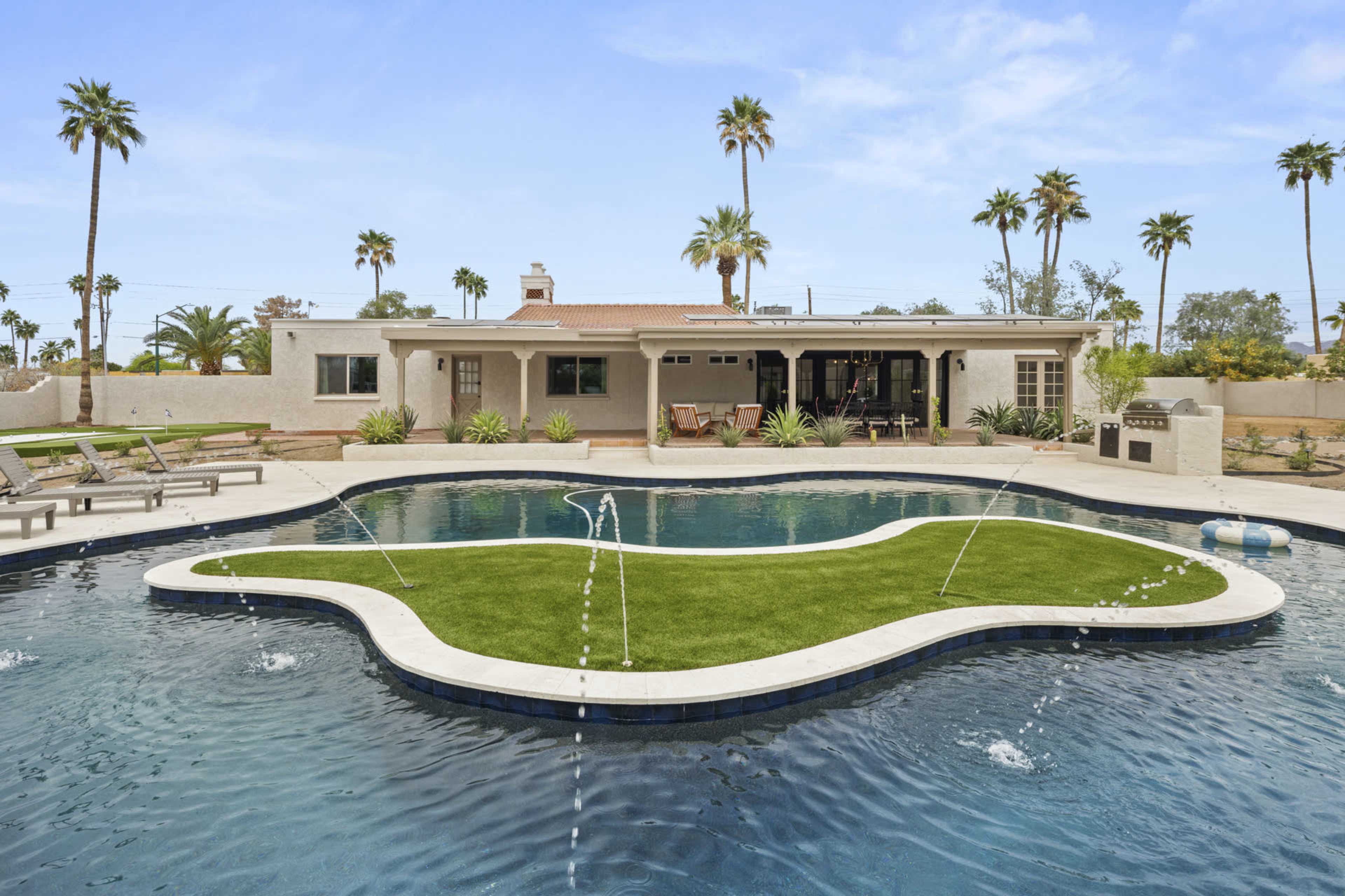 A modern backyard features a swimming pool with an island of artificial grass in the center, surrounded by lounge chairs and palm trees.