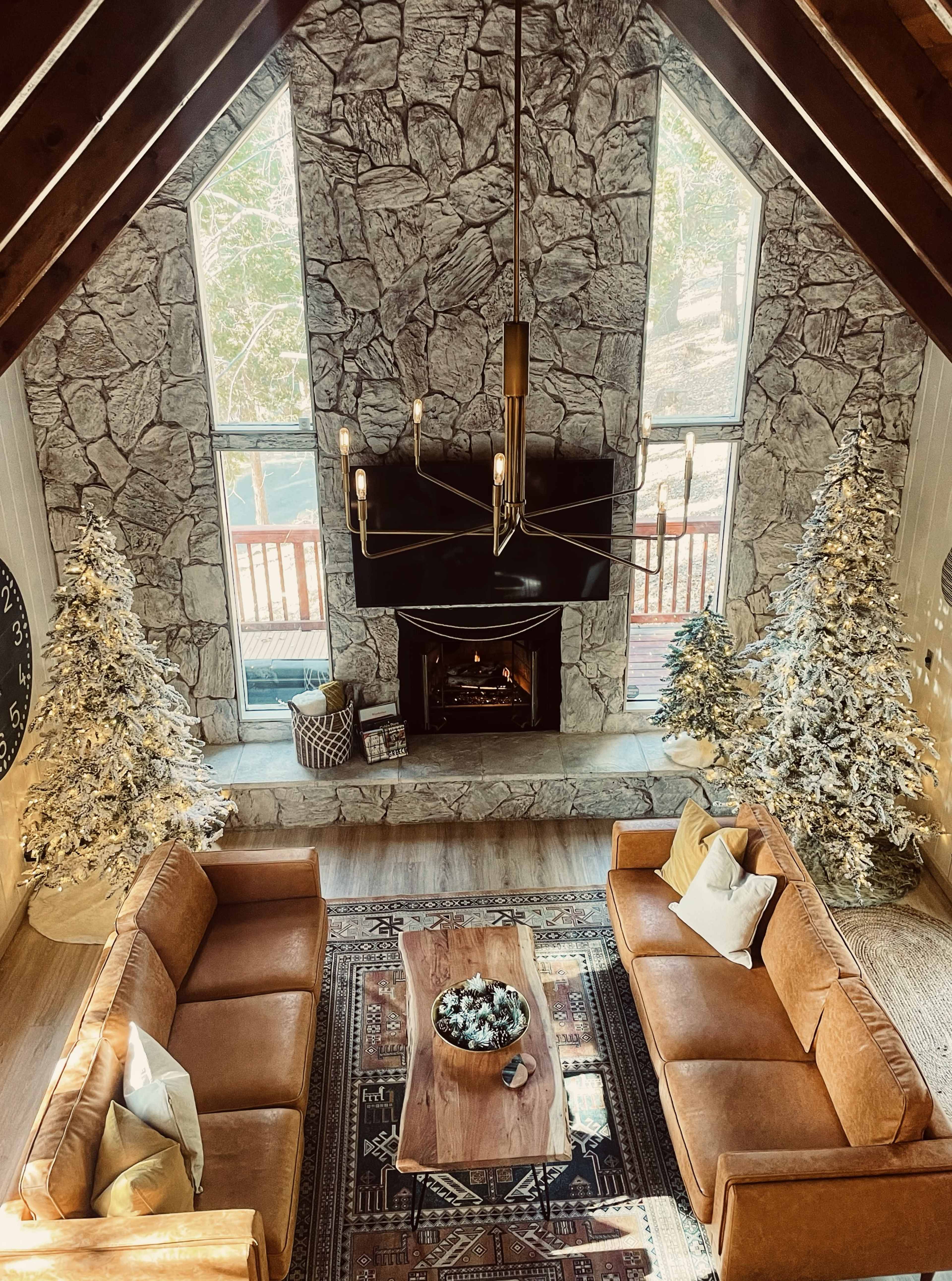 The image shows a spacious living room with a stone fireplace, two brown sofas facing a wooden coffee table, and two decorated Christmas trees flanking the fireplace.