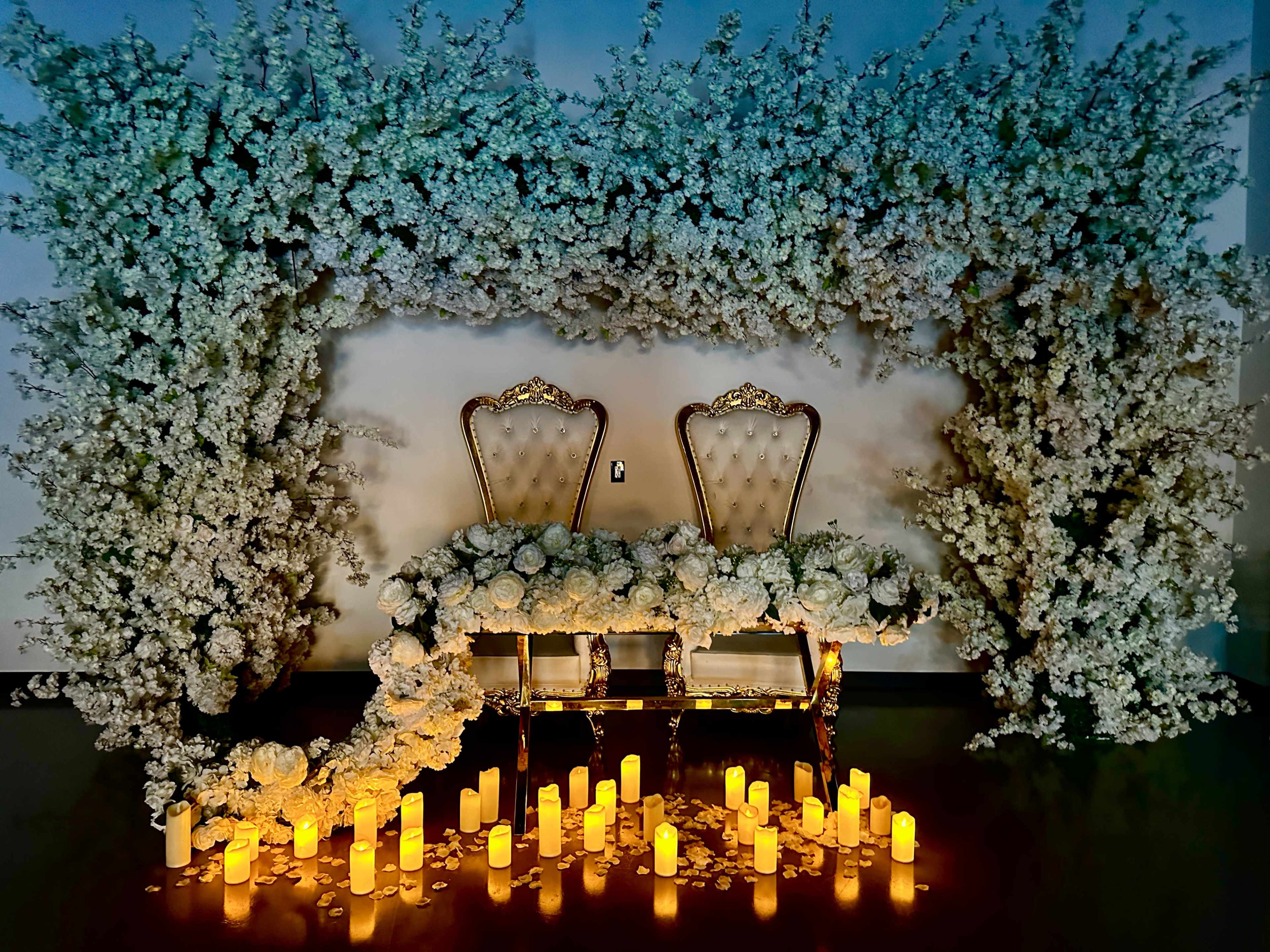 An elaborately decorated backdrop with two ornate chairs surrounded by artificial floral arrangements and lit candles on the floor.