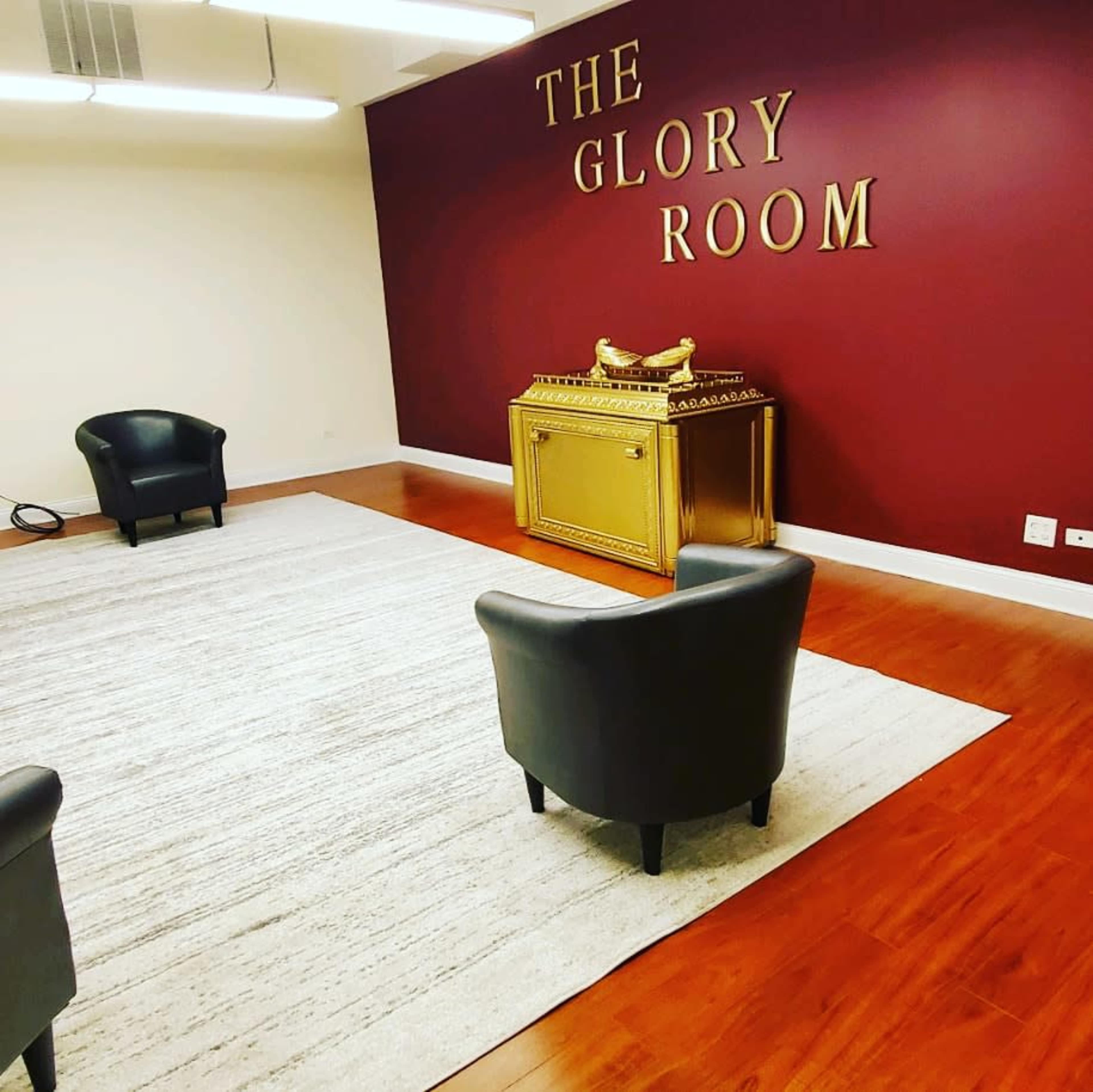 A room labeled "The Glory Room" with two black chairs, a decorative golden chest, and a light-colored area rug on a wooden floor.