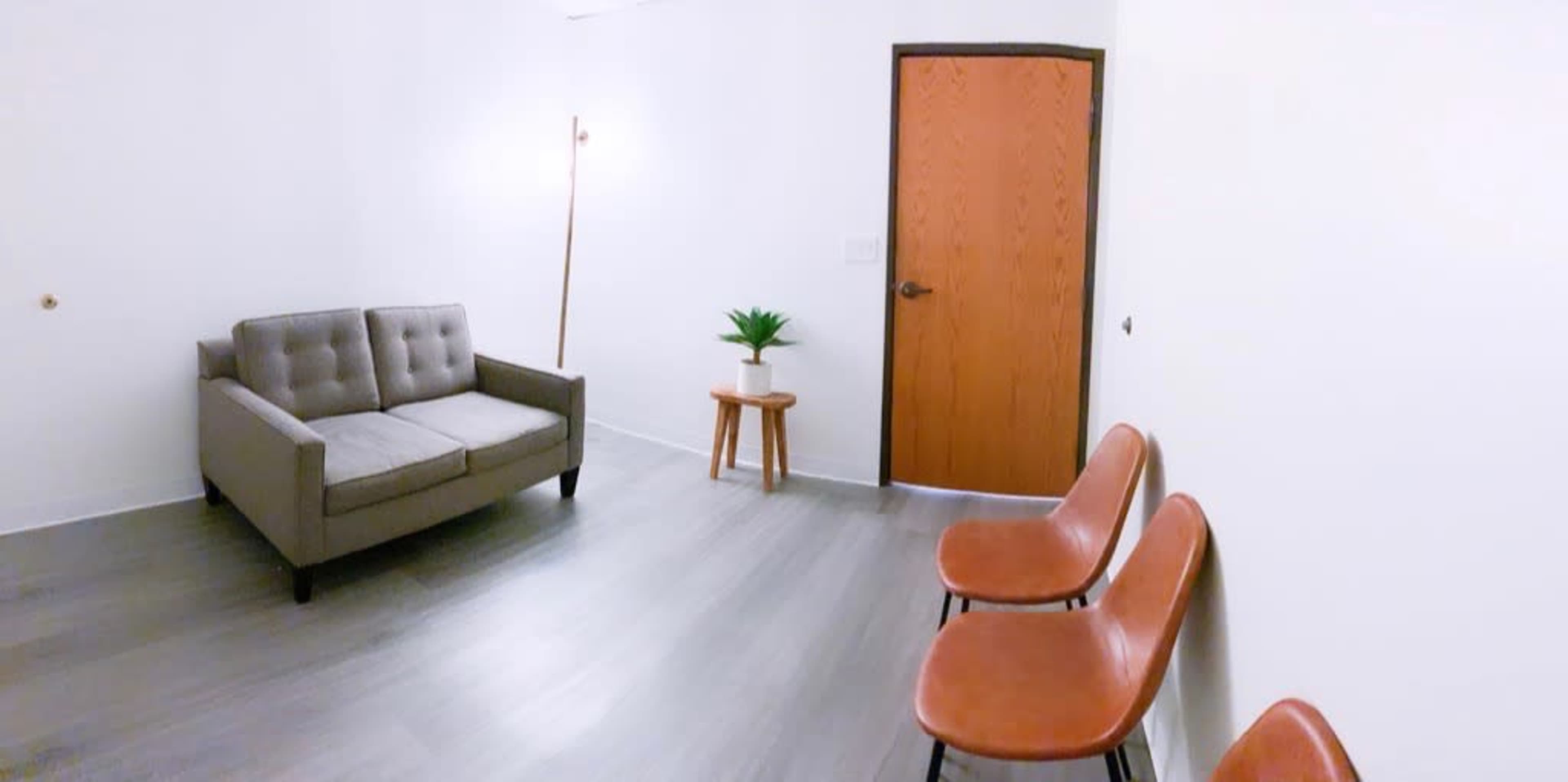 A small room with a gray sofa, two brown chairs, a potted plant on a wooden side table, and a wooden door.
