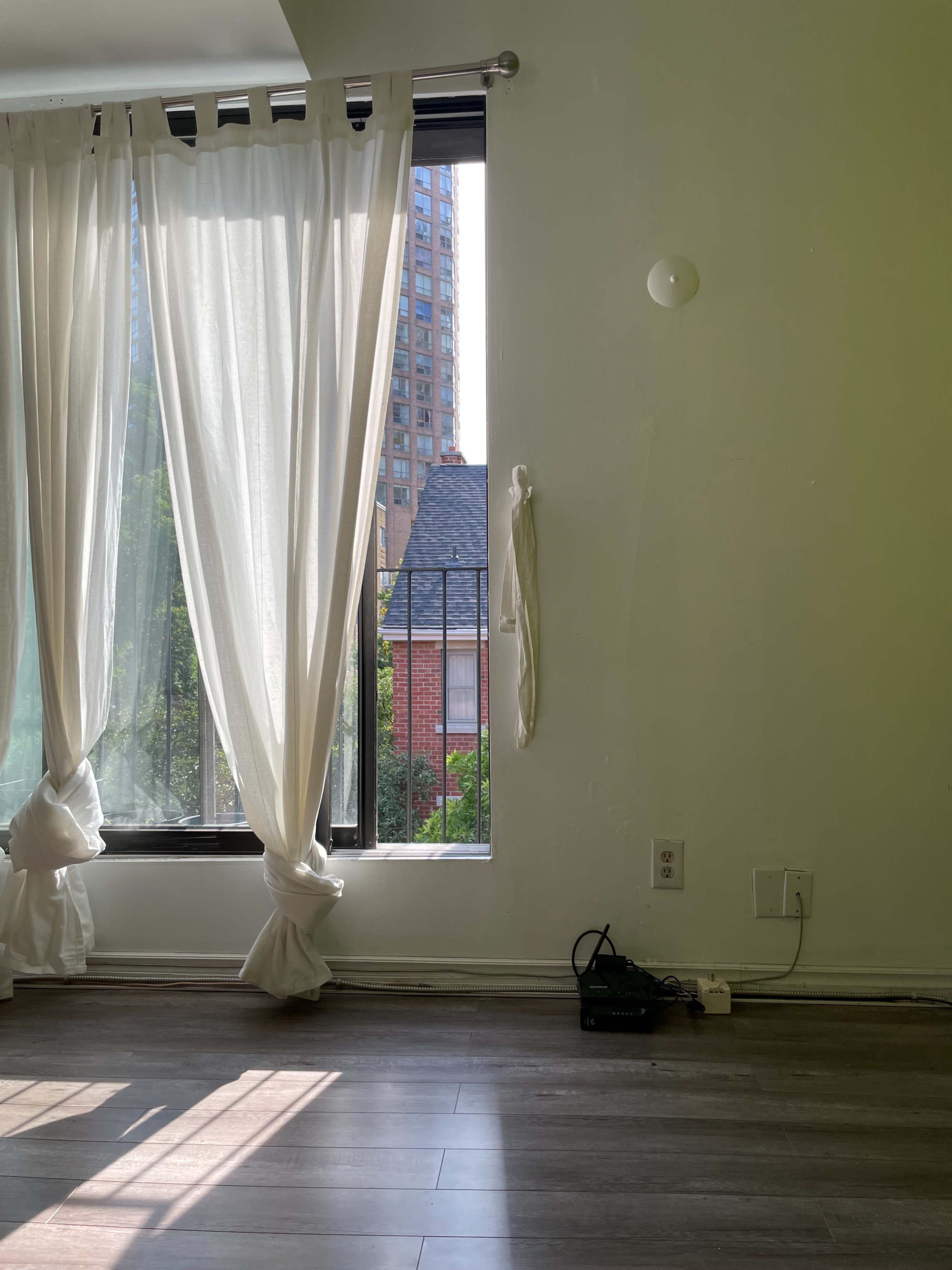 A sunlit room features sheer white curtains framing a window that overlooks a cityscape, with a small black device on the floor nearby.