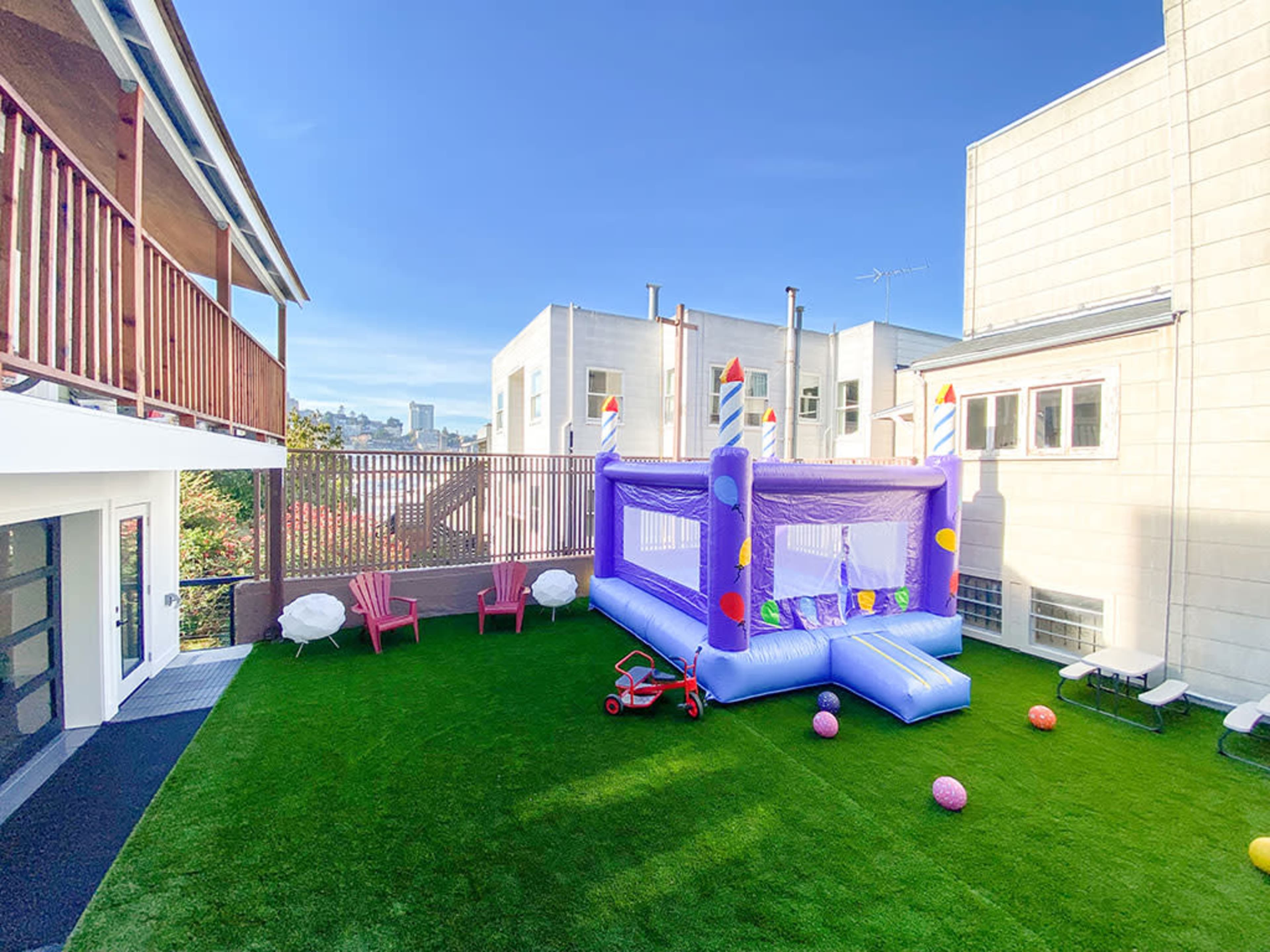 A colorful inflatable bounce house set up in a neatly maintained backyard with artificial grass, surrounded by lounging chairs and playground balls.