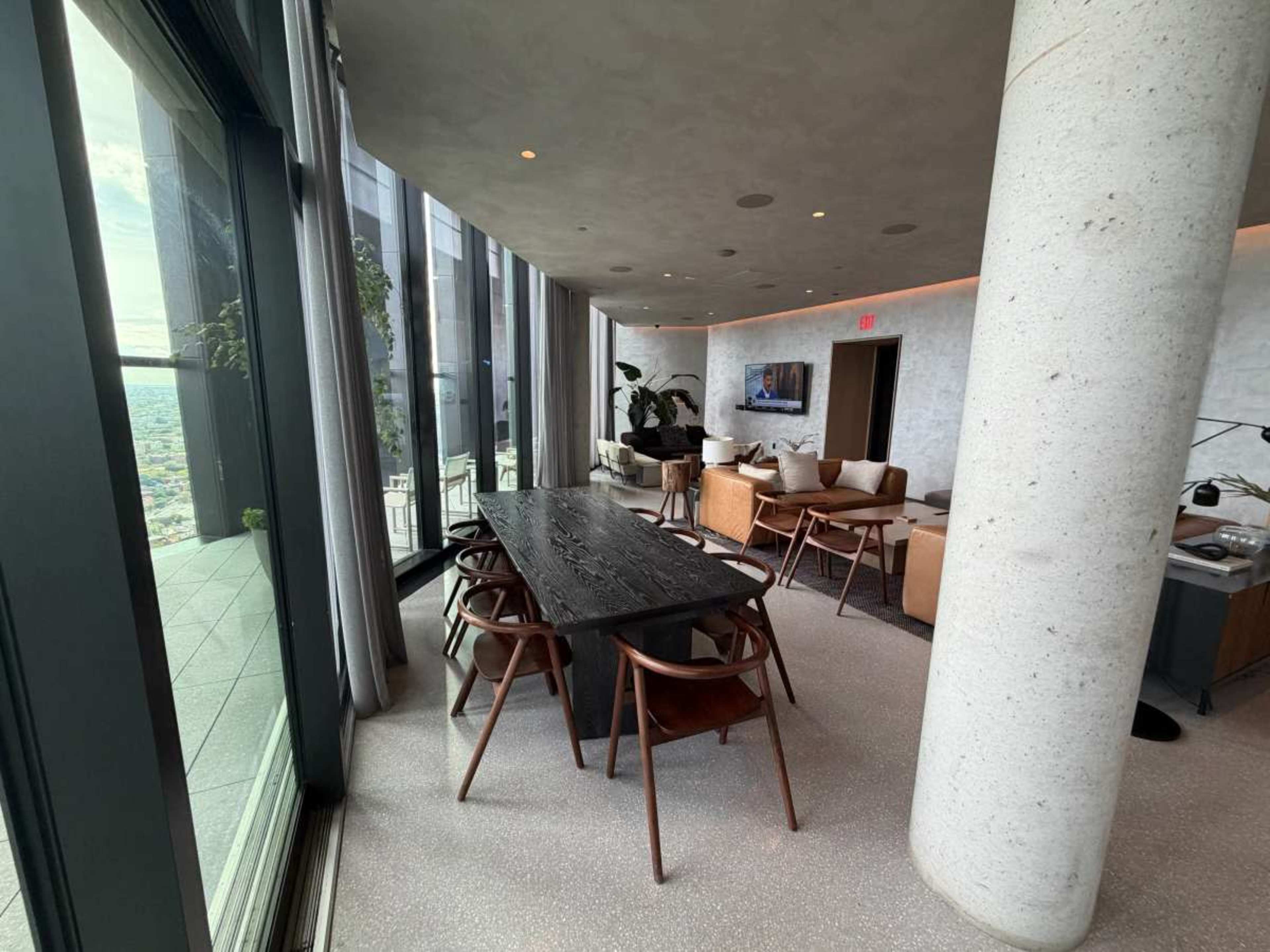 The image shows a modern dining area with a long black table surrounded by wooden chairs, large windows offering a view of the city, and a comfortable seating area with light-colored furniture.