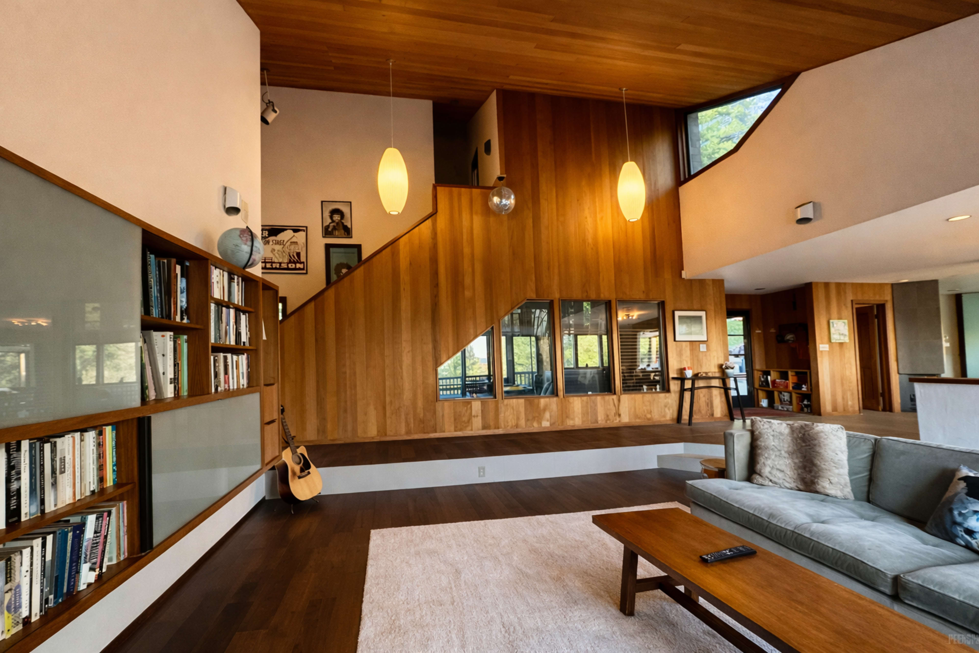 The image shows a contemporary living room with a wooden staircase, large windows, and a mix of bookshelves and seating areas.