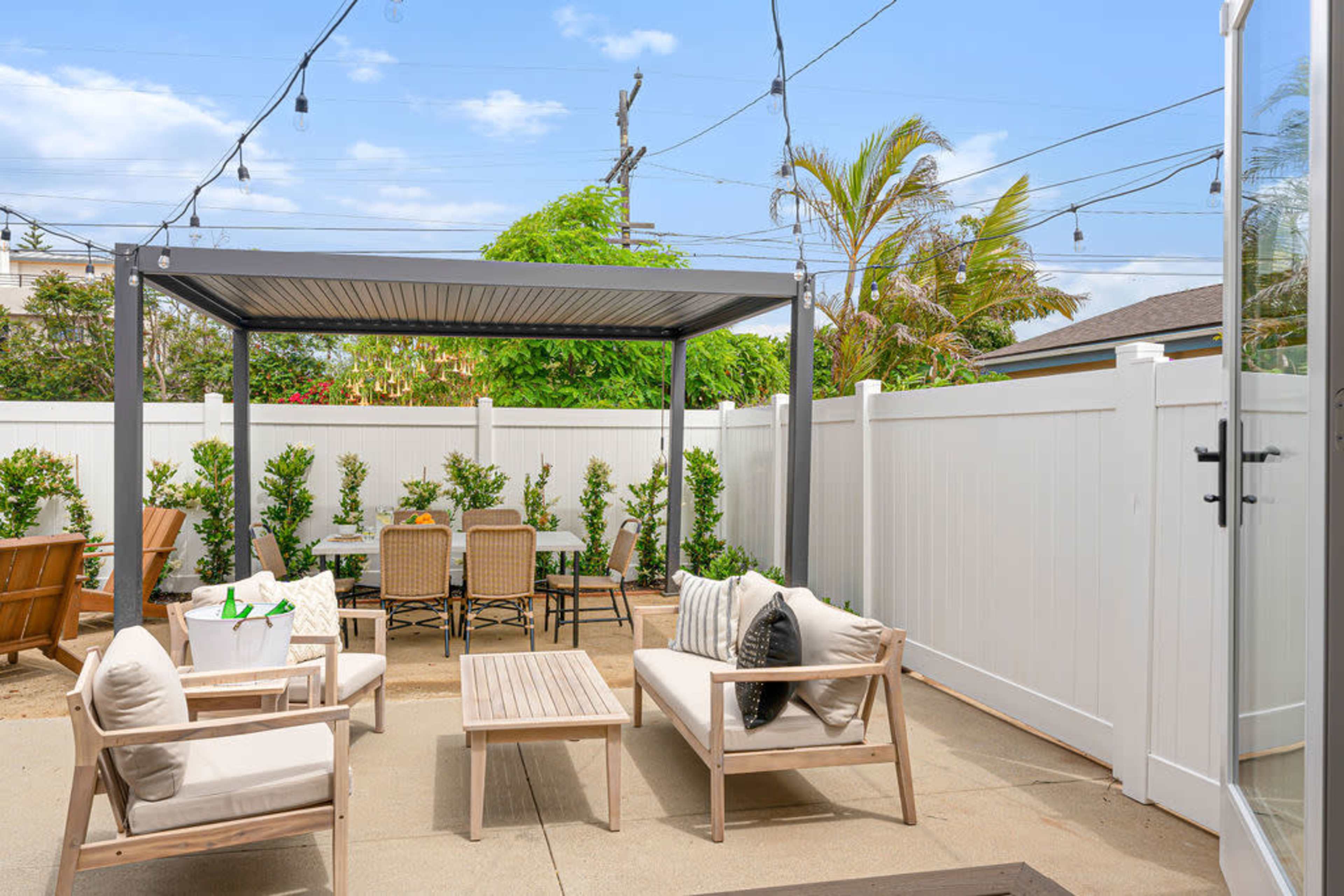 The image shows a patio area with a pergola, outdoor seating, a dining table, and greenery against a white fence.