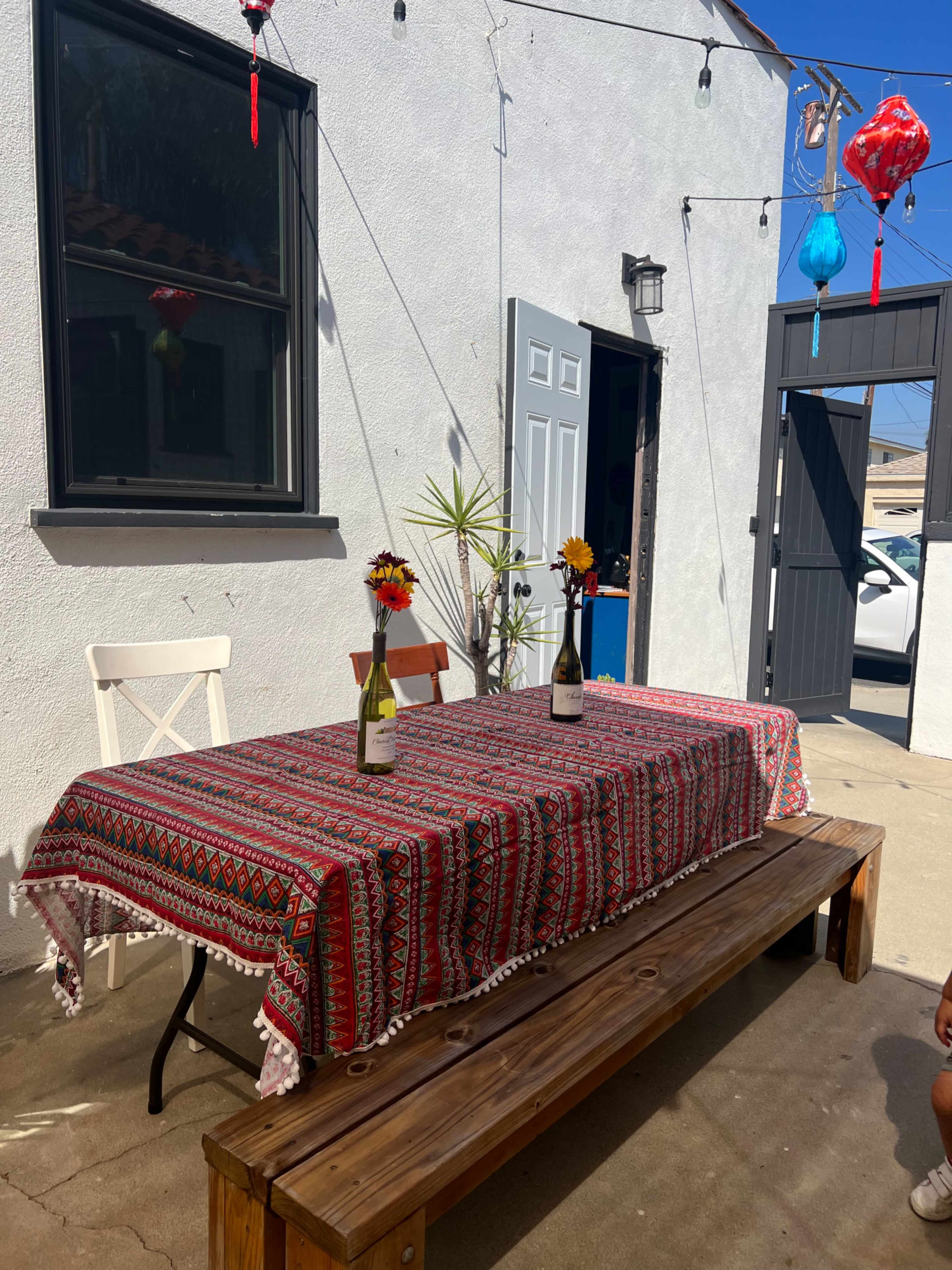 A table with a patterned tablecloth and two bottles sits outdoors in front of a light-colored wall, flanked by a blue door and a plant.