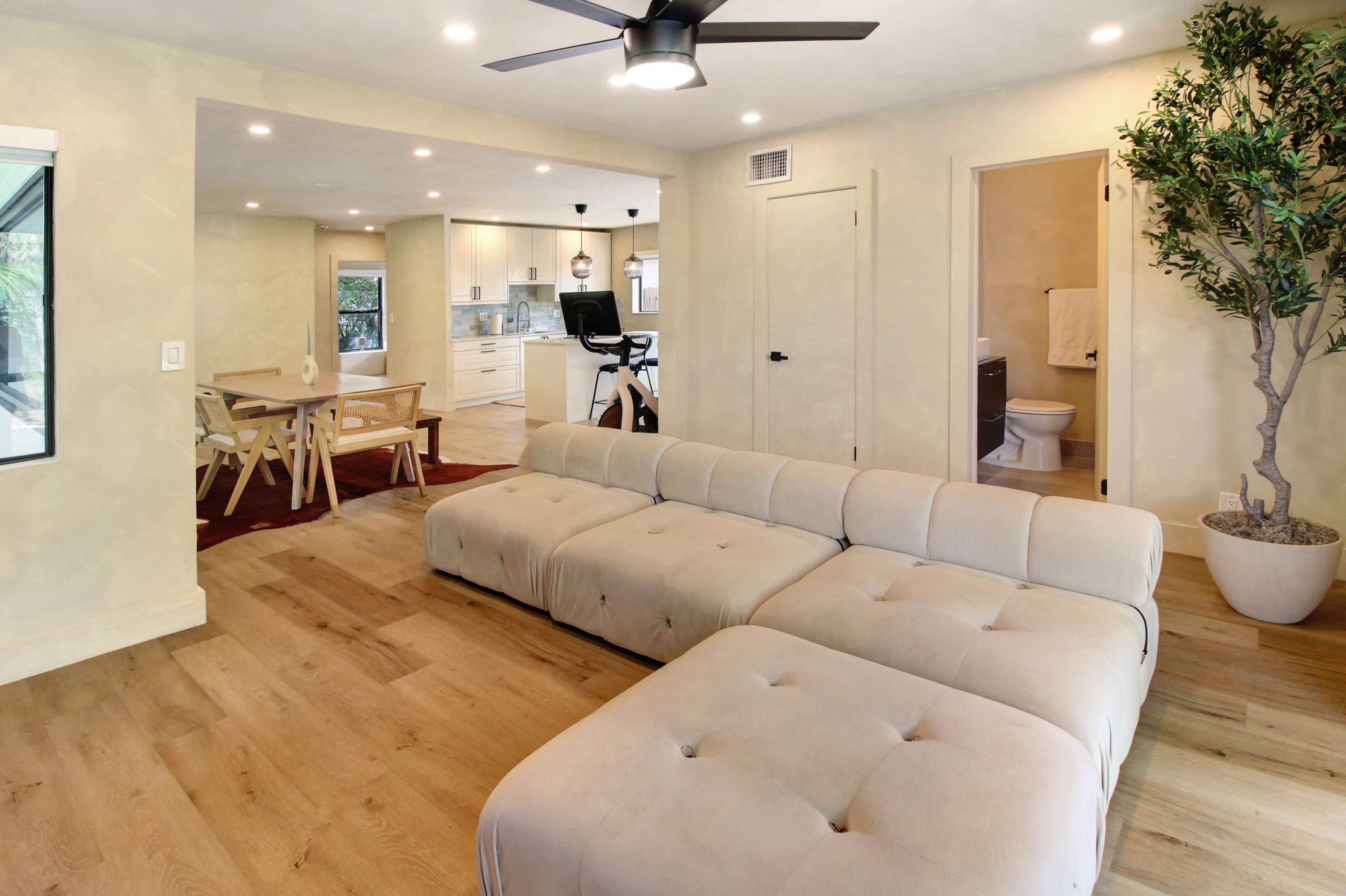 The image shows a modern living room featuring a large beige sectional sofa, a wooden dining table, and an open kitchen area with bright lighting.