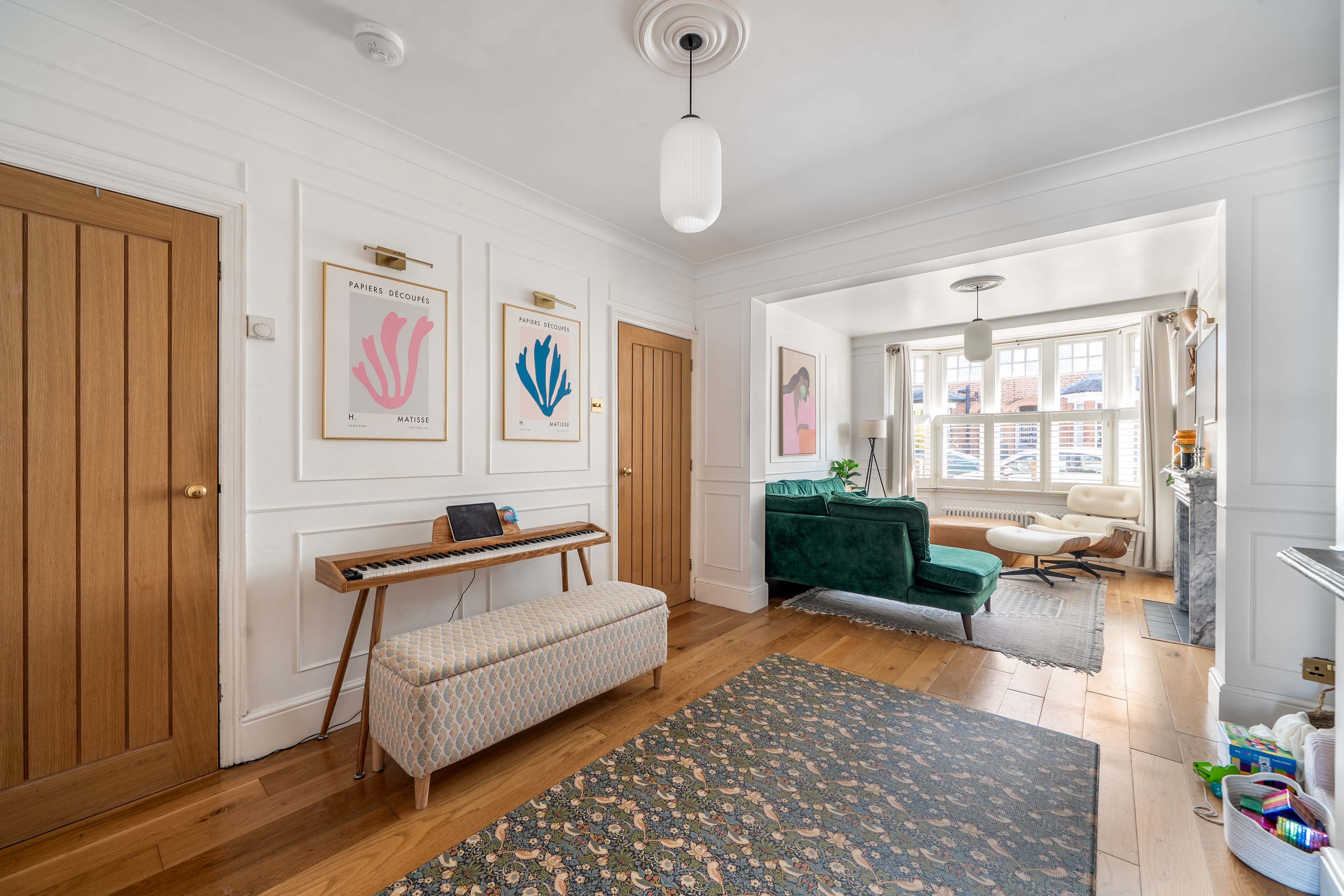 The image shows a bright interior living space featuring a decorative rug, a vintage keyboard on a bench, and framed artworks on the walls, with a green sofa and an armchair visible in the adjacent area.