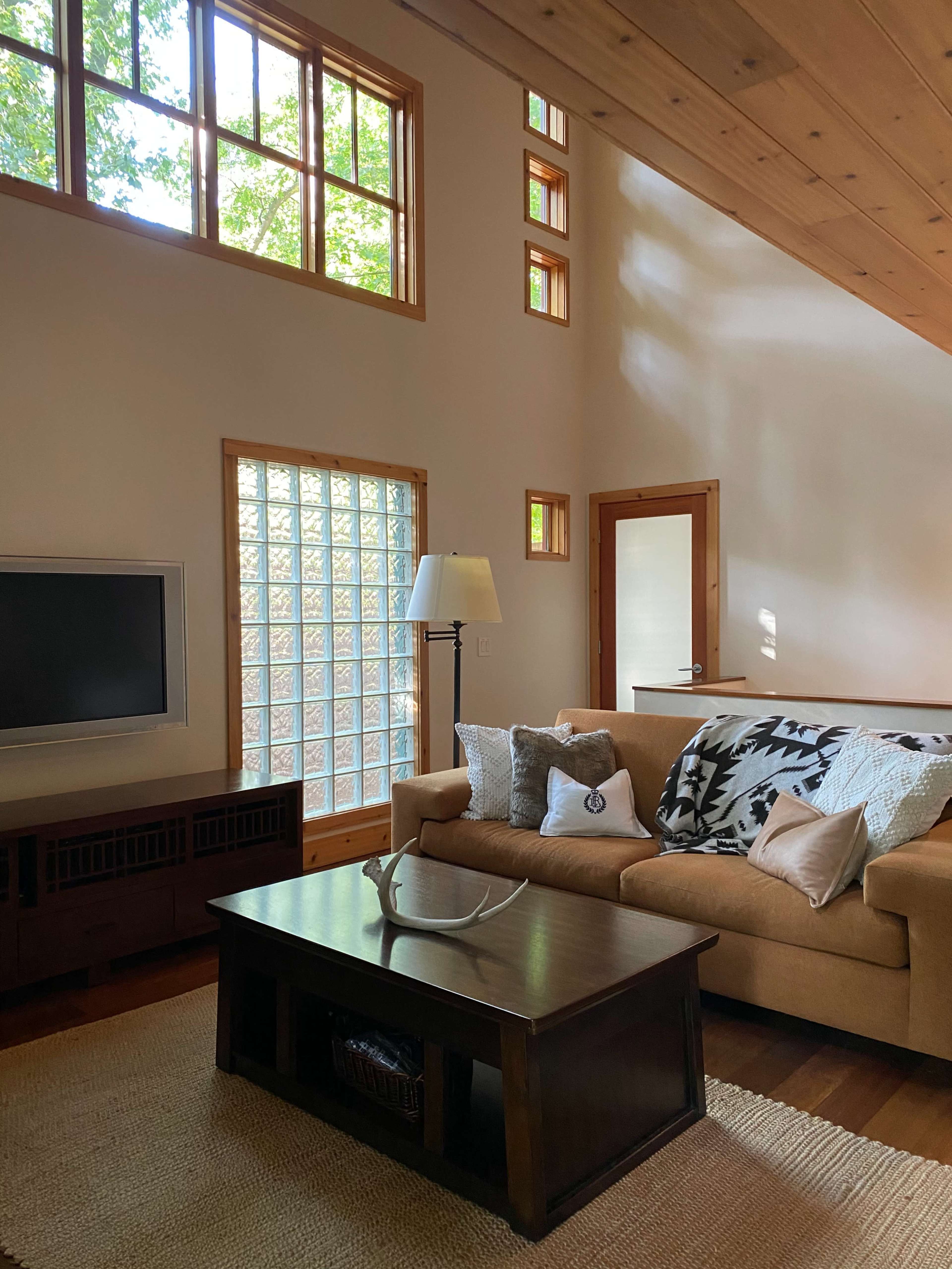 A cozy living room features a tan sofa with decorative pillows, a wooden coffee table, a television, and large windows allowing natural light to fill the space.