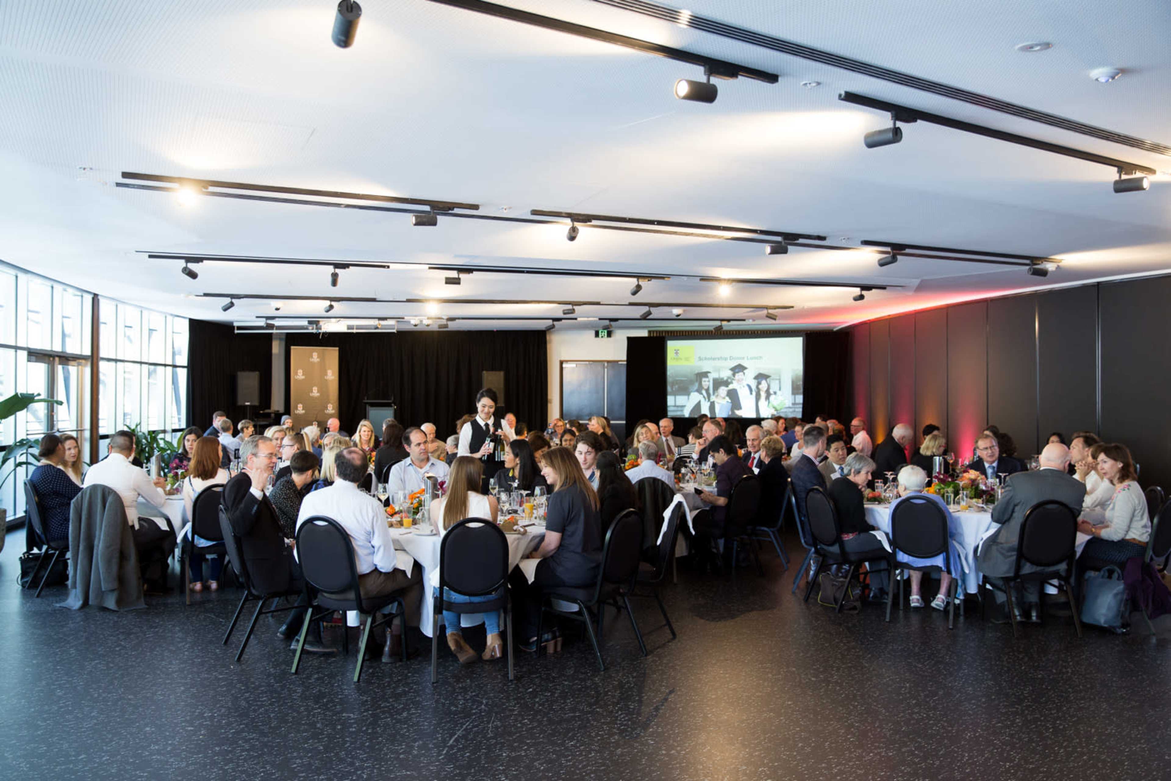A large group of people is seated around multiple tables in a spacious, well-lit event hall, enjoying a meal together.