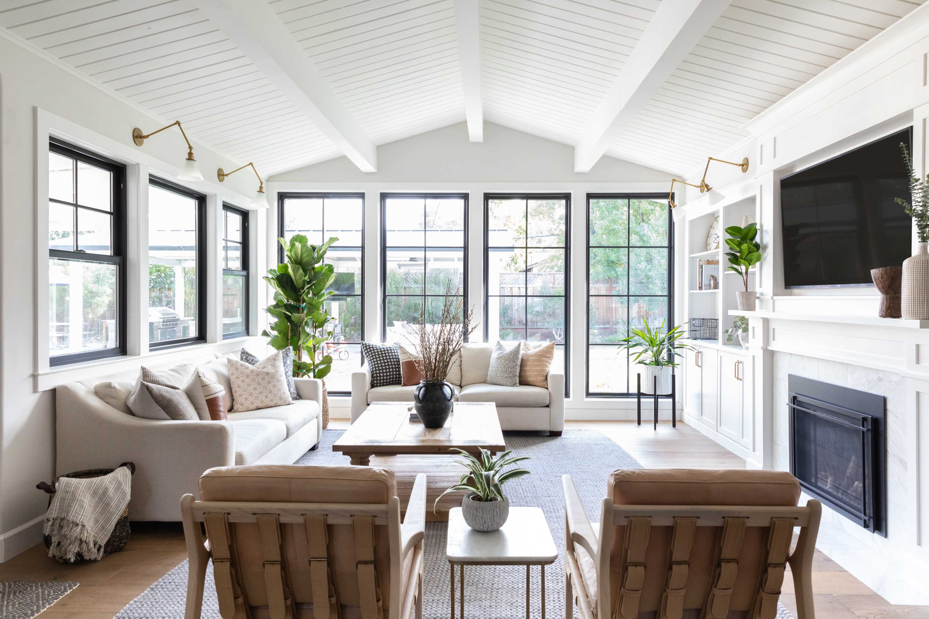 A bright living room features a white sofa, wooden chairs, and large windows with views of greenery.