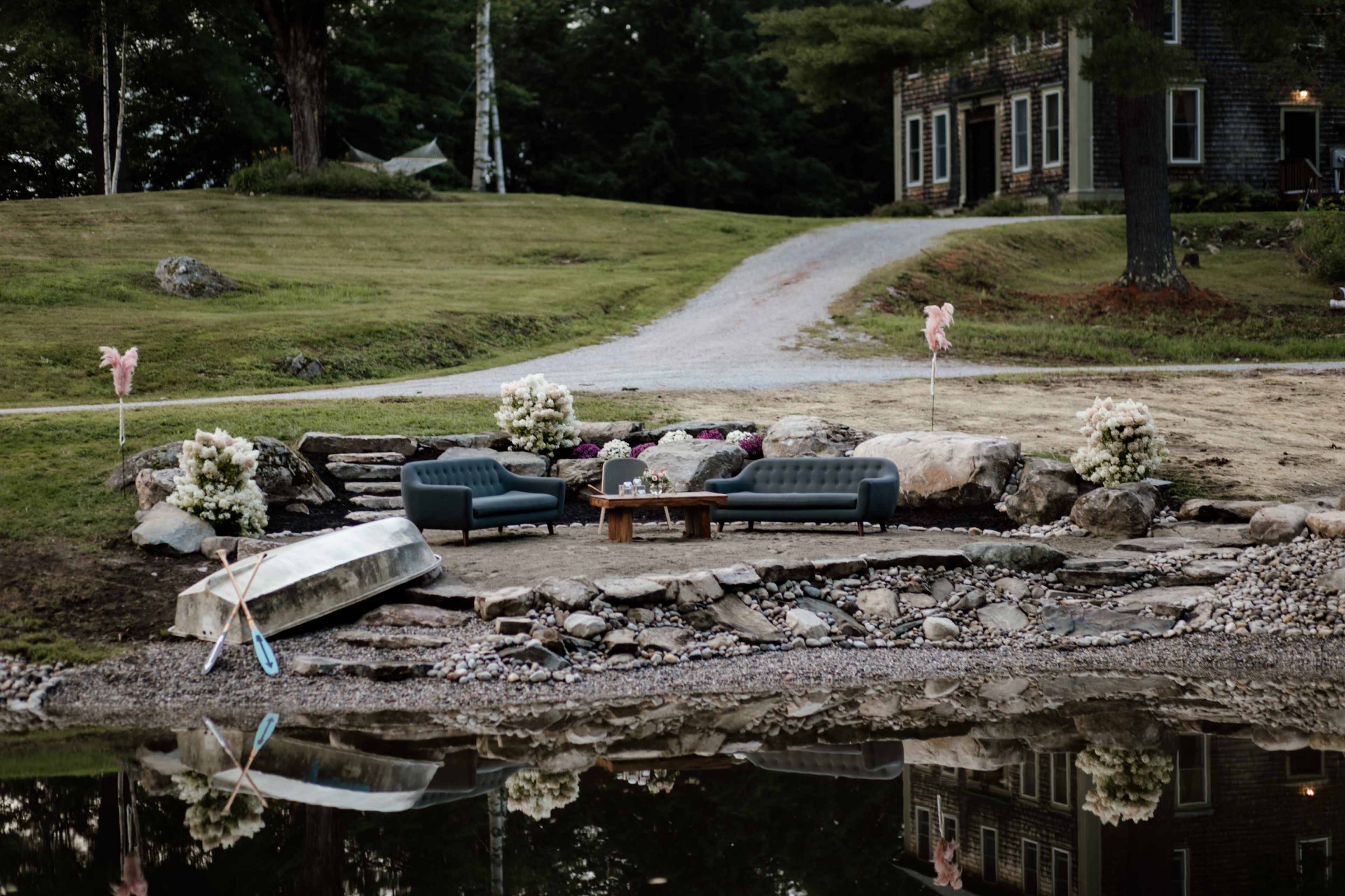 A seating area with two sofas, a table, and flower arrangements is set by the edge of a lake, with a gravel path leading to a house in the background.