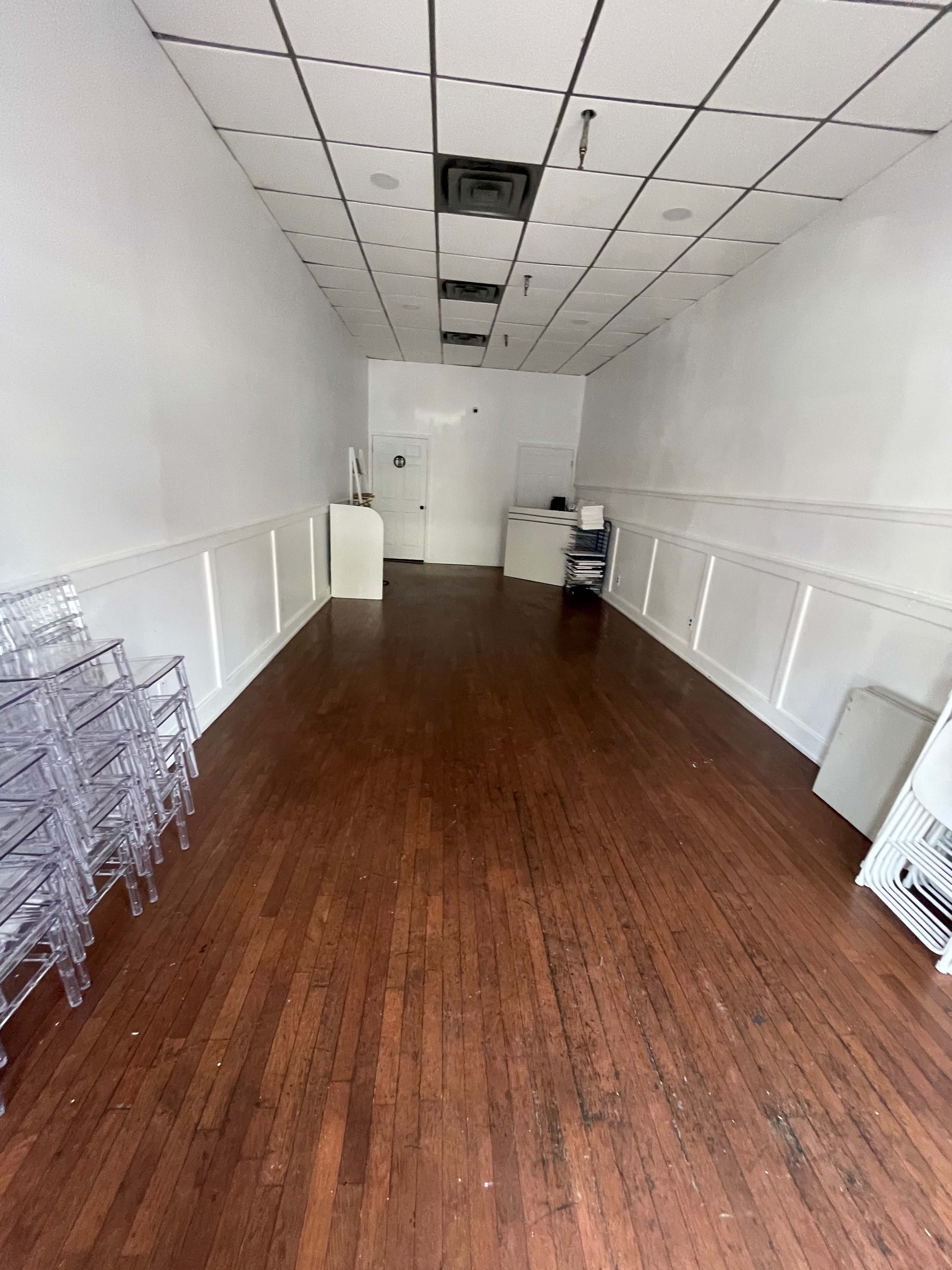 A vacant room with wooden floors, white walls, and minimal furnishings, including some clear chairs stacked against one side.