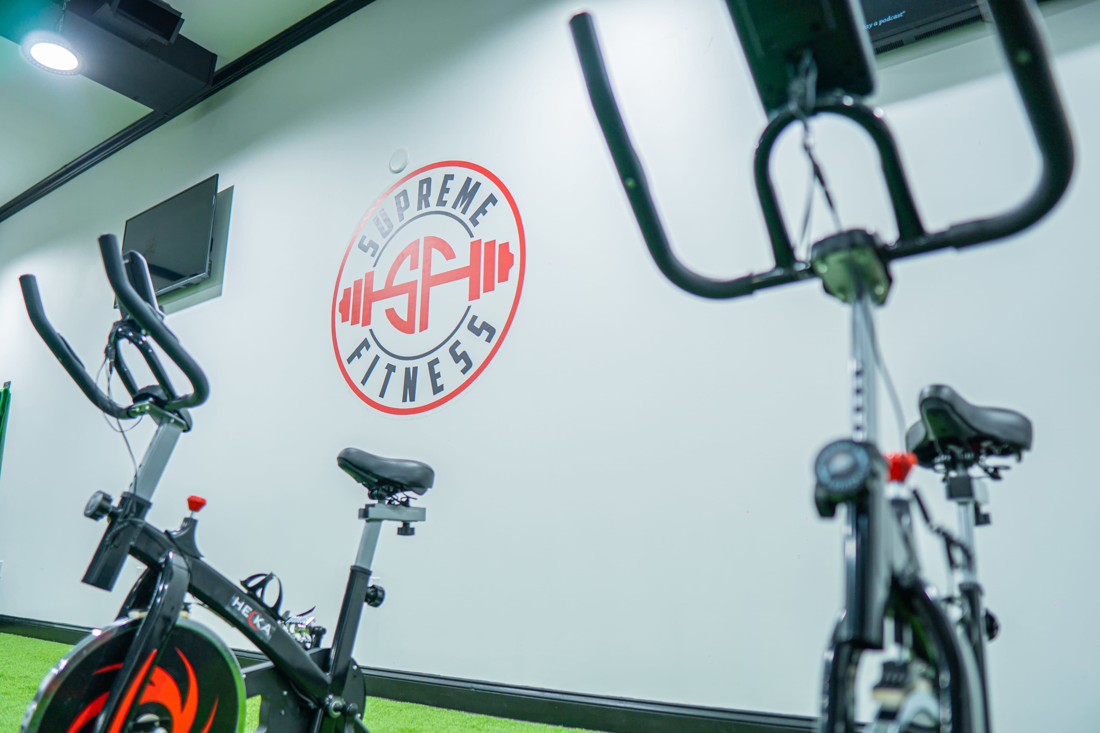 Two stationary exercise bikes positioned in front of a "Supreme Fitness" logo on a wall.