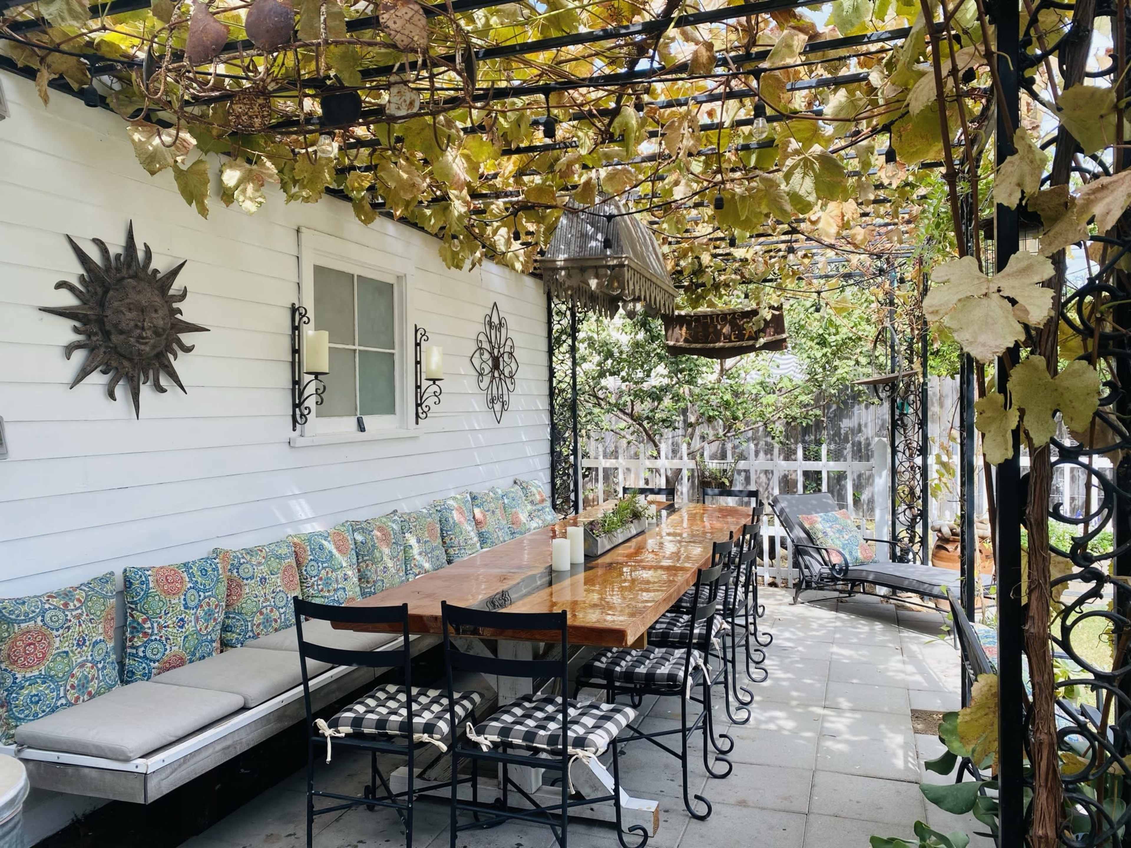 A long wooden table surrounded by black metal chairs is set under a vine-covered pergola on a patio.