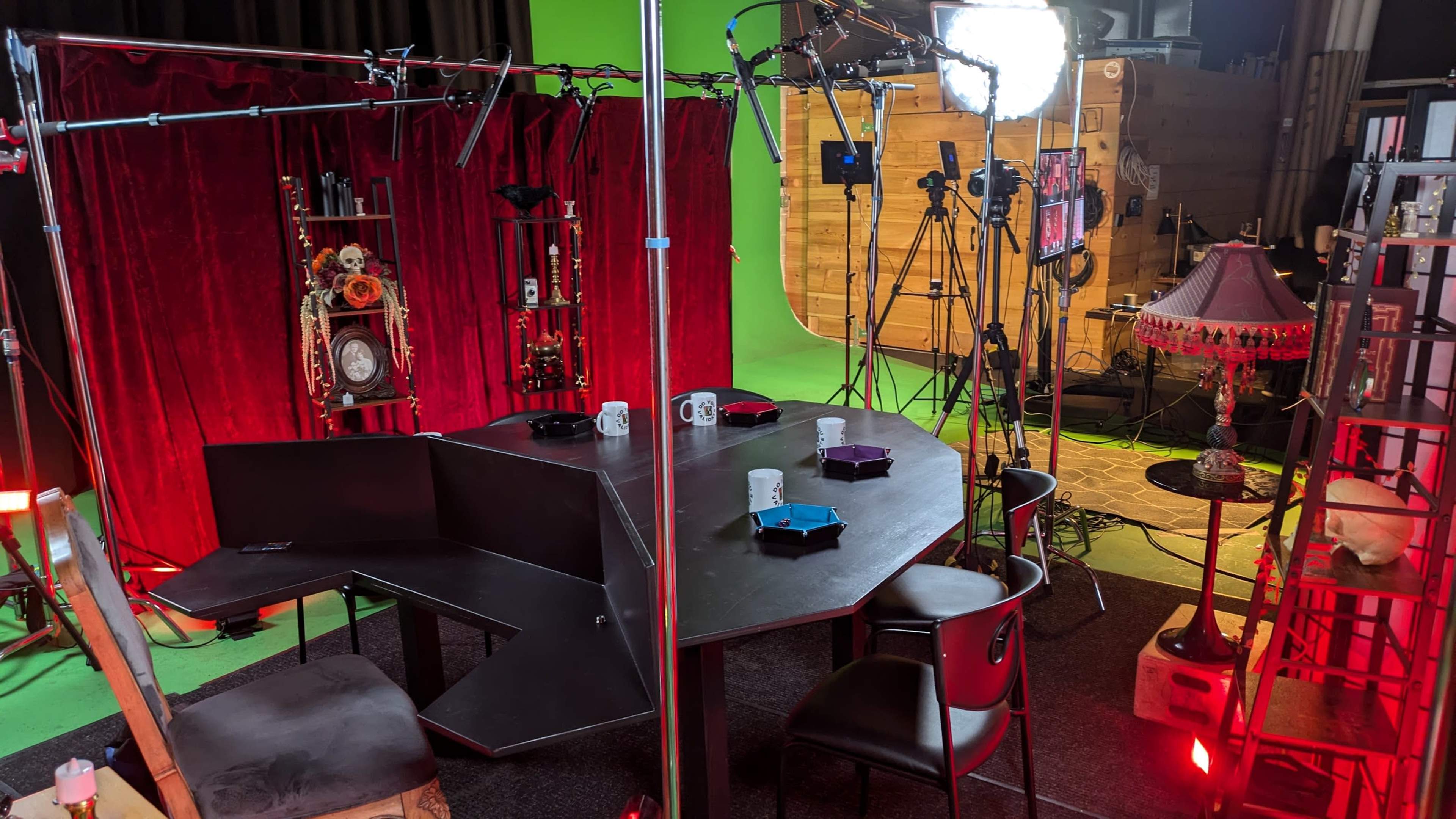 The image shows a film set featuring a round table surrounded by chairs, with a backdrop of red curtains and various lighting and camera equipment positioned around the scene.