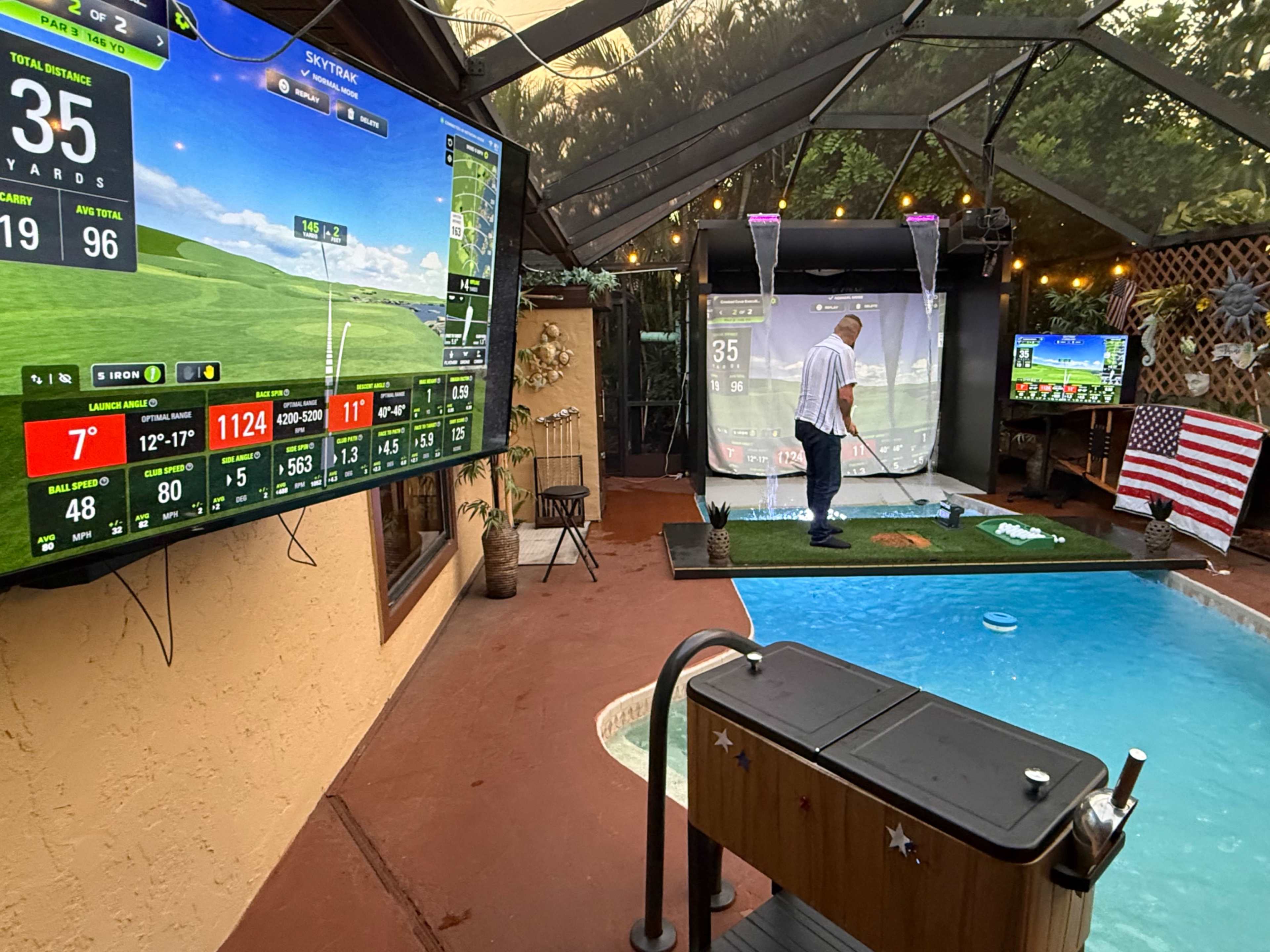 A golfer practices on a putting green inside a screened patio with multiple large display monitors showing golf statistics and a golf simulator.