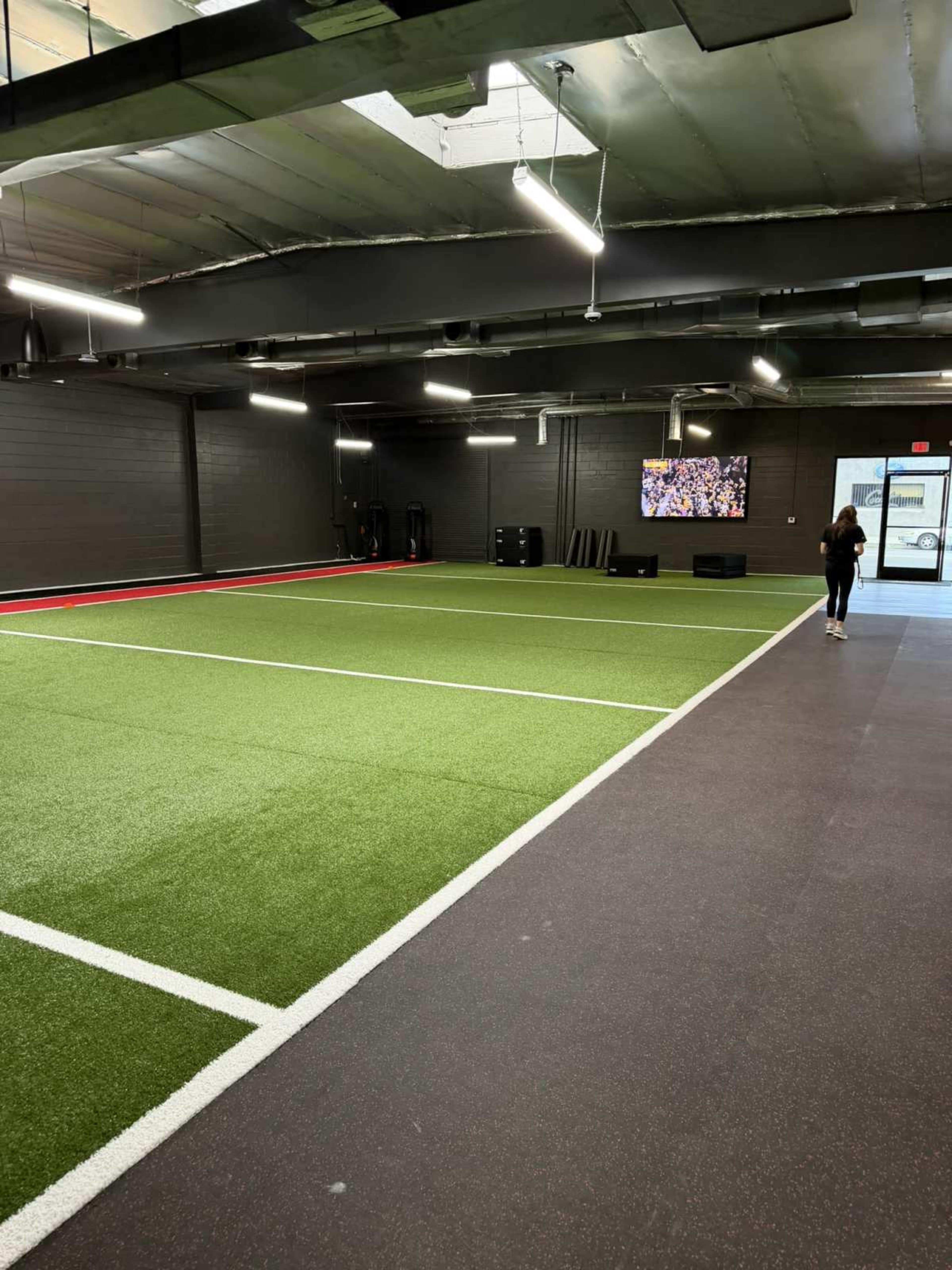 The image shows a spacious indoor athletic facility featuring green turf flooring, marked with white lines, and a television mounted on the wall.