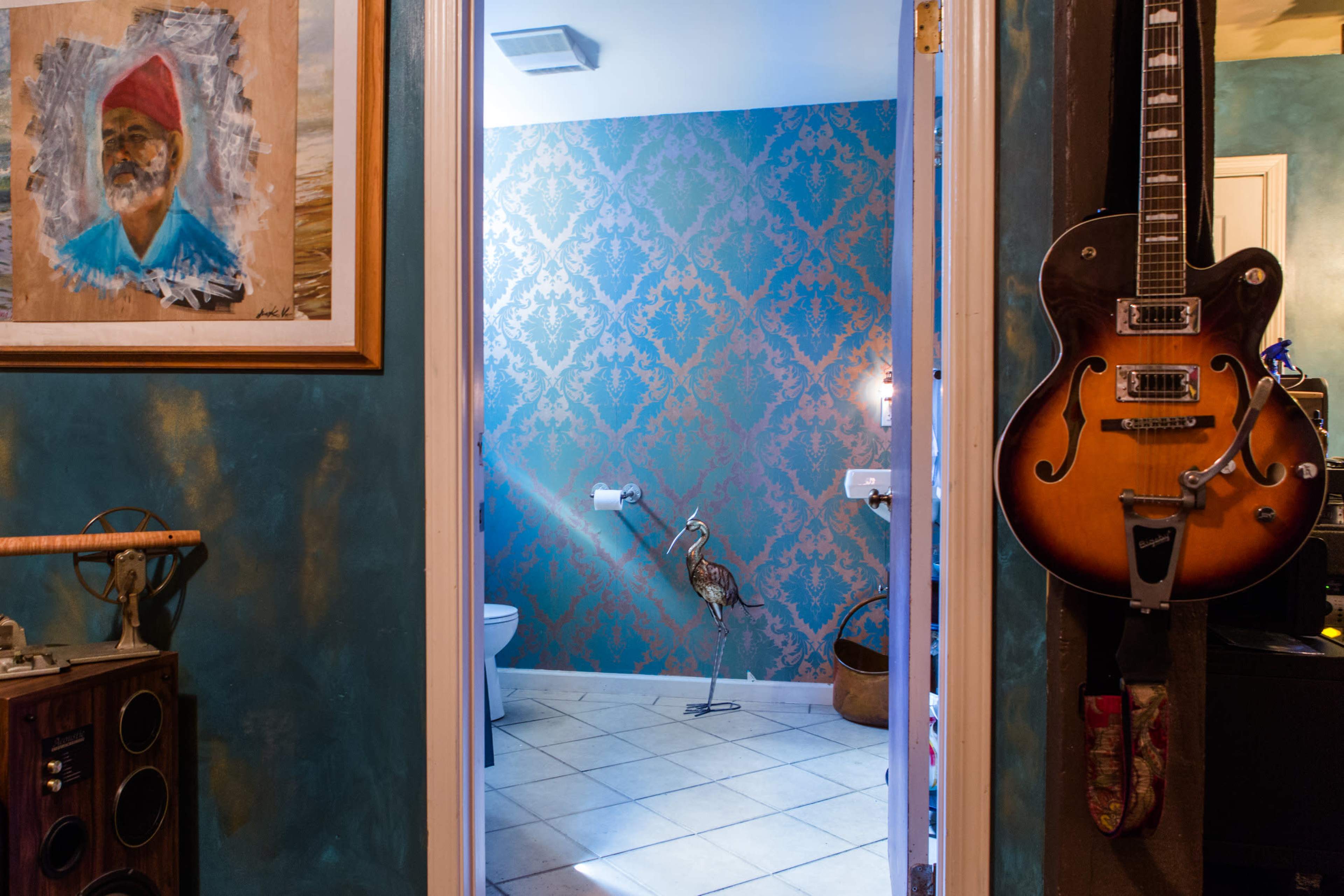 A brightly colored bathroom with a decorative wall, a toilet, and a crane statue is visible through a door, framed by a guitar and an artwork on the adjacent wall.