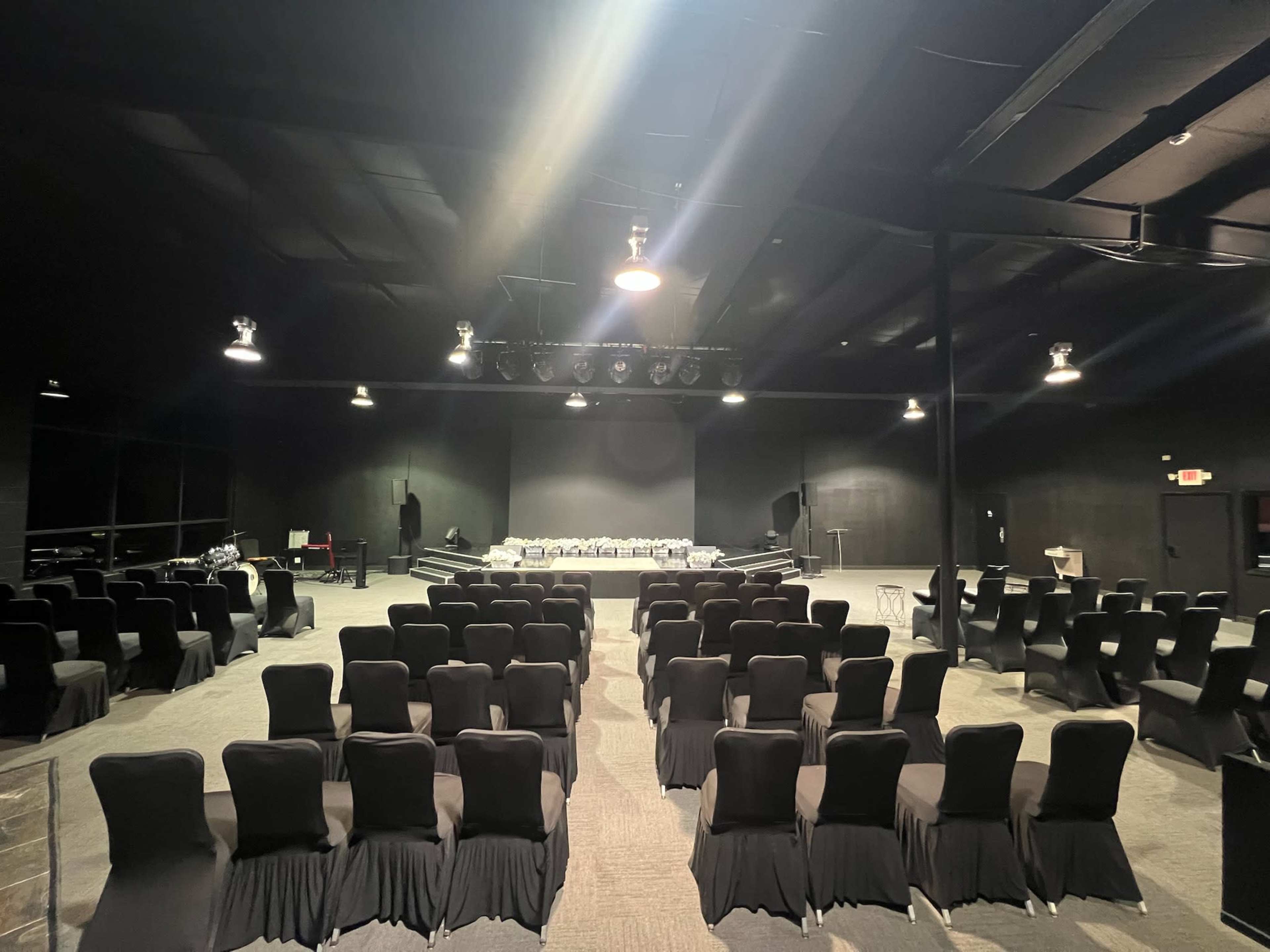 A large, dark room is set up with rows of black chairs facing a stage, illuminated by overhead lights.