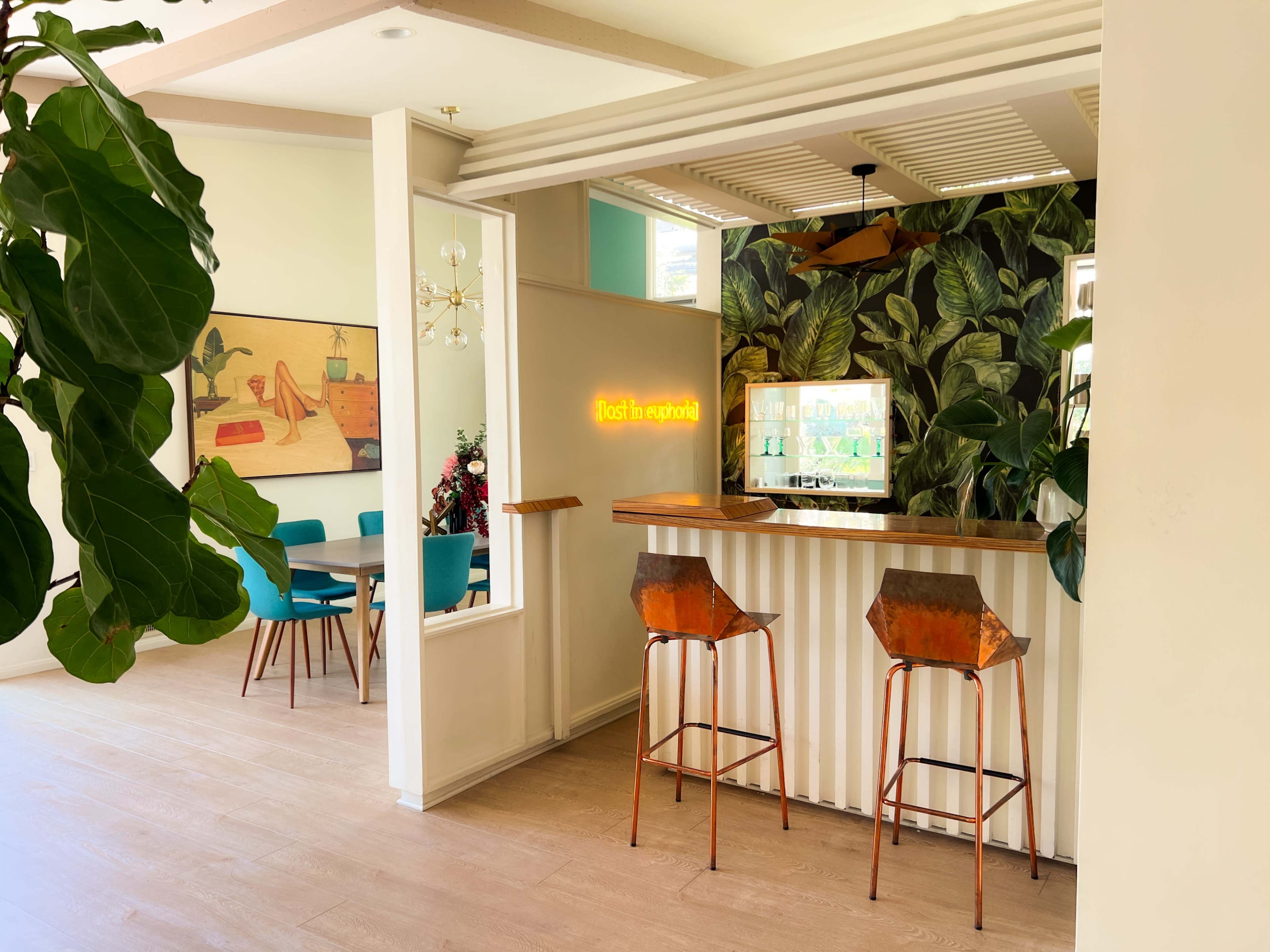 A modern interior space featuring a bar area with two copper stools, a decorative wall with plants, and a dining table in the background.